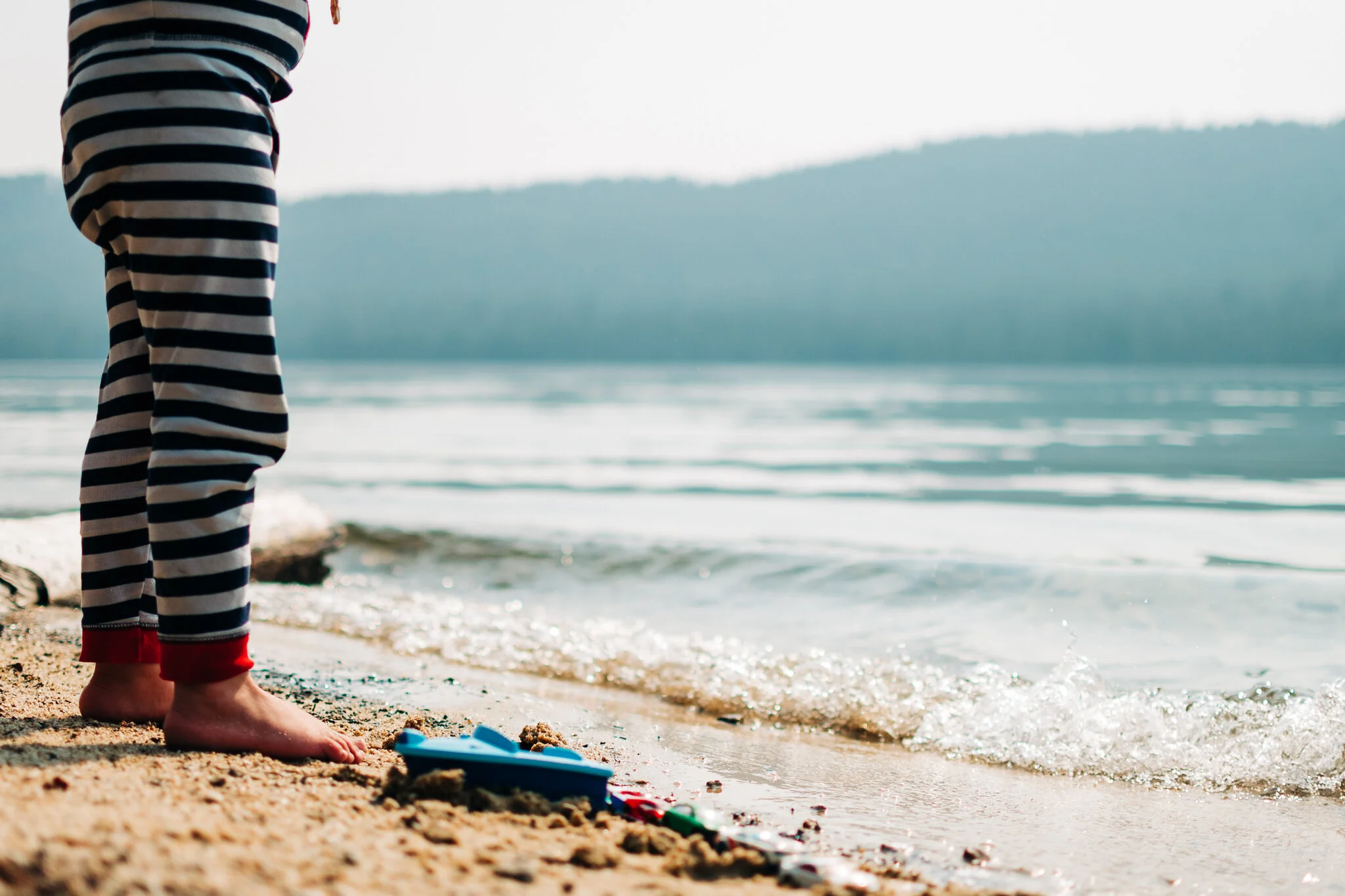 Child in striped pajamas standing at the shoreline with sand toys on the beach, water and mountain in the background.