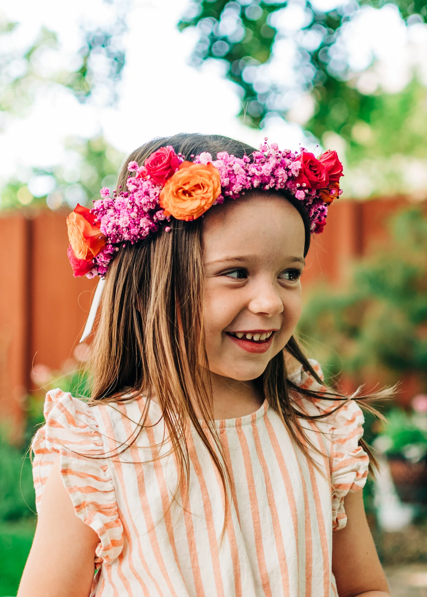 A young girl with brown hair wearing a floral crown made of pink, red, and orange flowers, smiling and looking to the side outdoors with a blurred background of trees and a wooden fence.