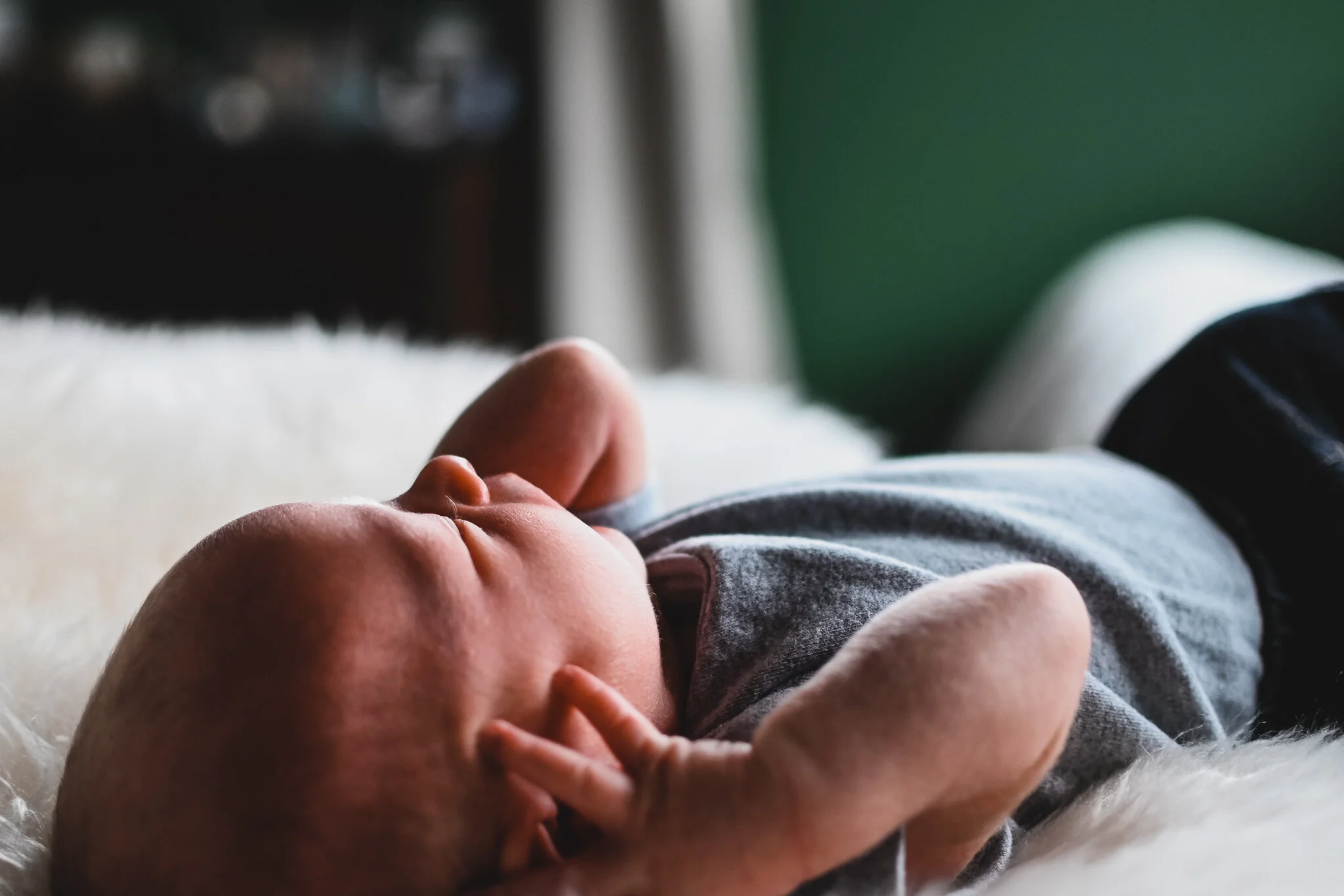 Close-up of a sleeping baby lying on a soft, white surface with one hand near their face, wearing a gray shirt.