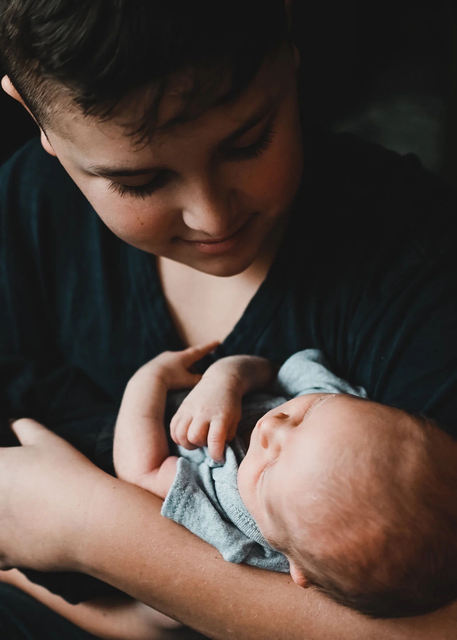 A young boy holds a newborn baby in his arms, looking down at the baby with a gentle expression.