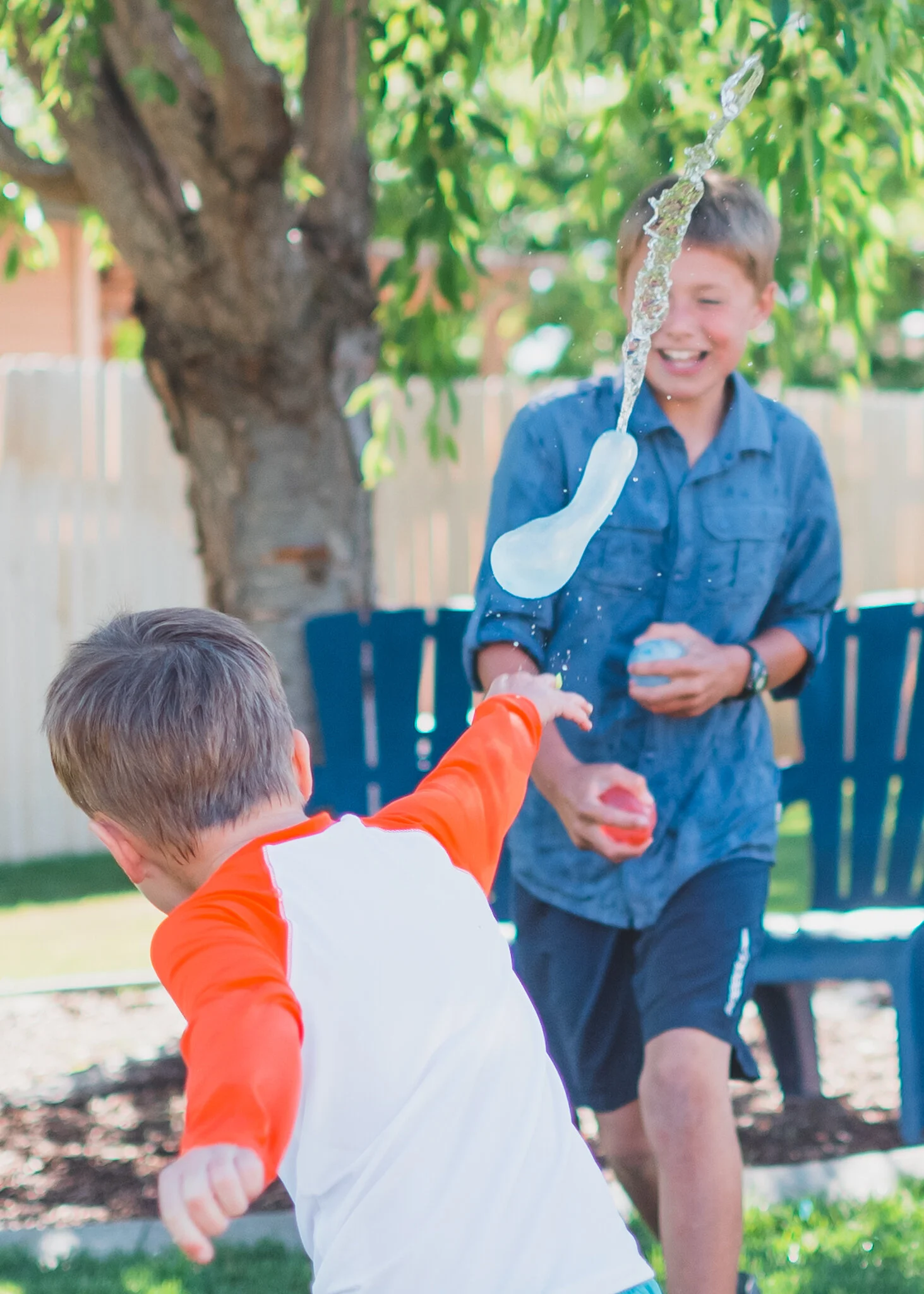 Two boys playing with a water gun outdoors, with a tree and a bench in the background.
