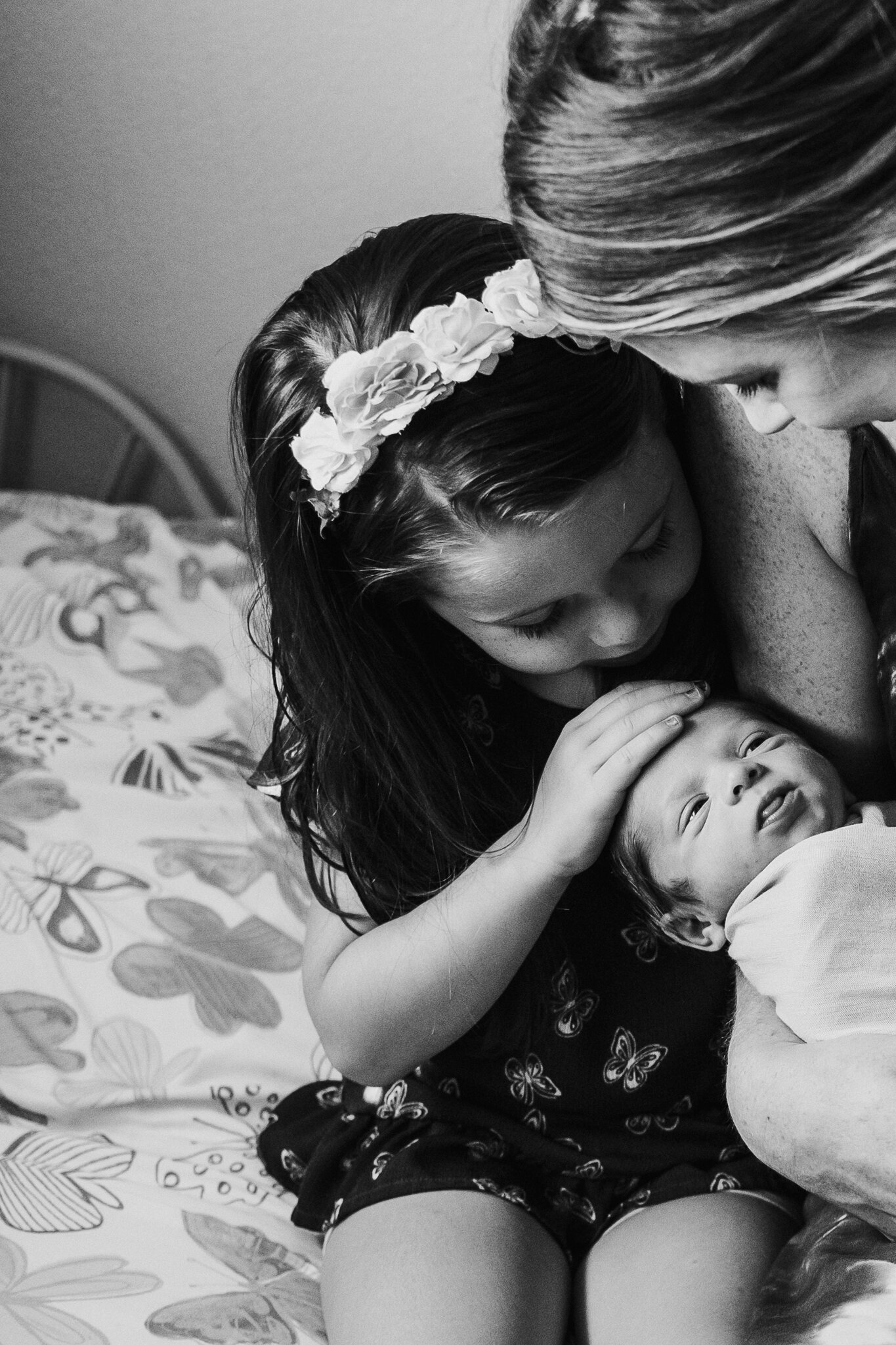 A woman with long dark hair and a floral headband is gently kissing a baby lying on her lap, while a woman with short hair is leaning in close, touching the baby's face in an intimate moment.