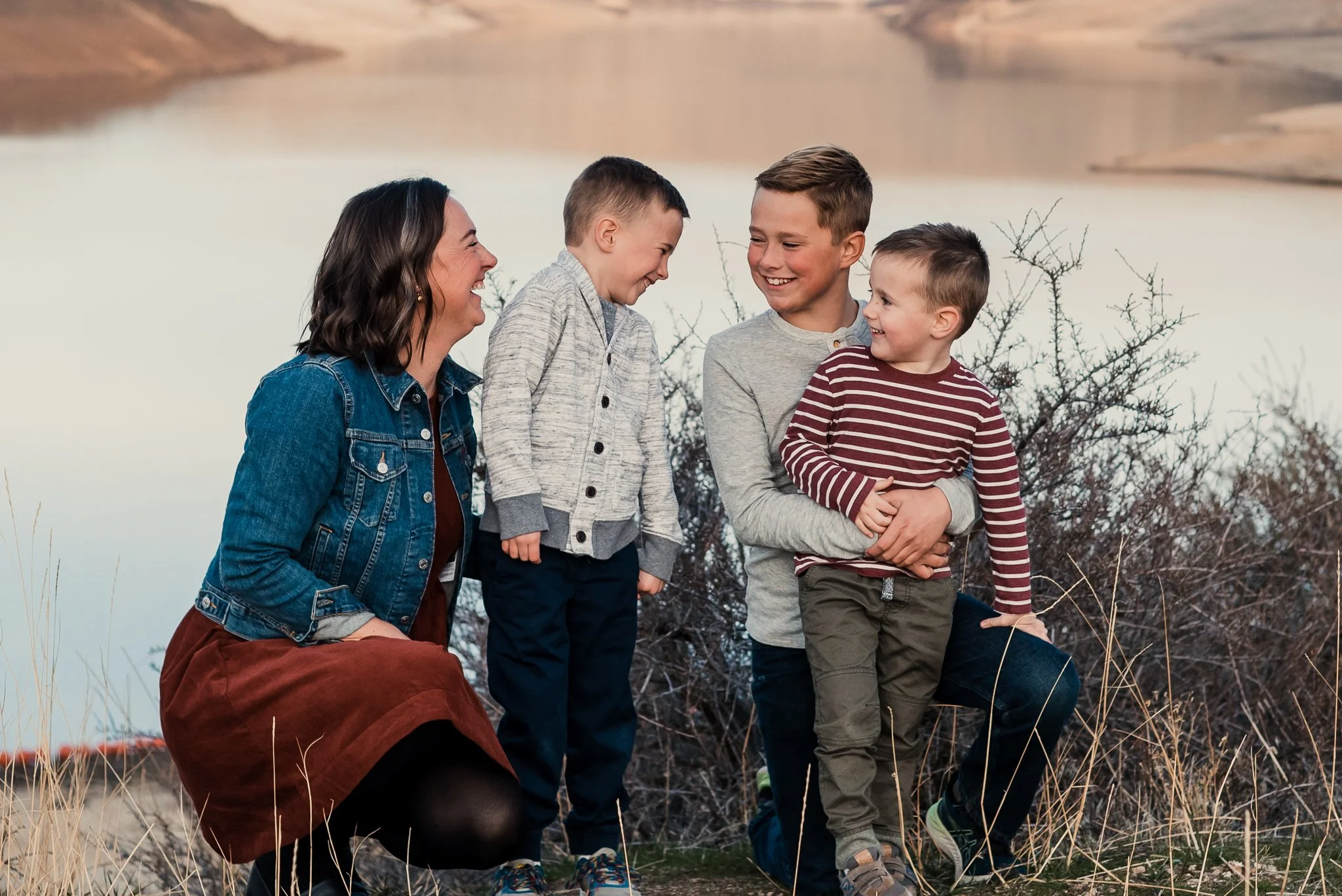 A woman and three boys smiling and laughing outdoors near a body of water with dry grass and bushes.