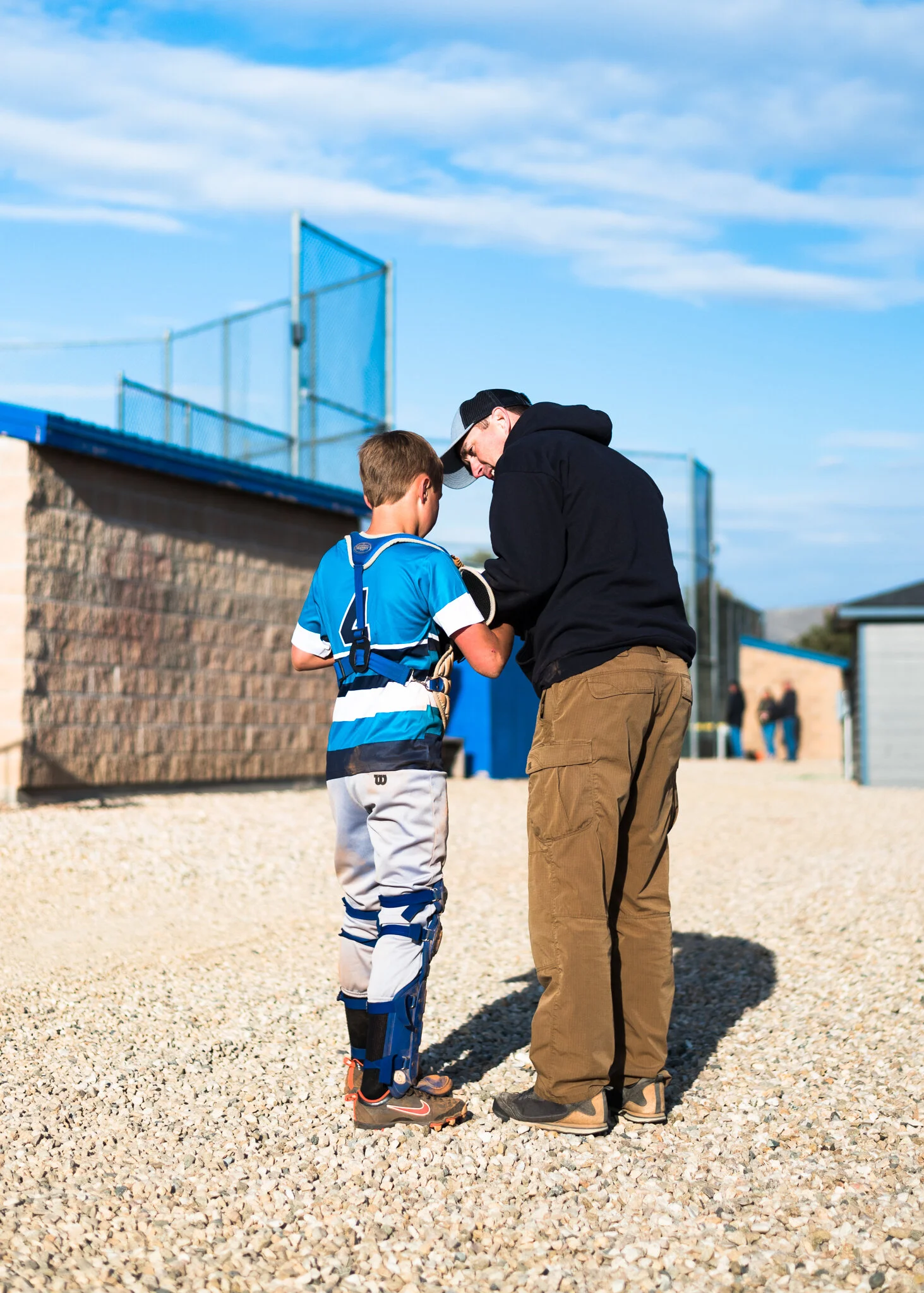 A young boy in baseball gear being fitted or given instructions by a man, possibly a coach or parent, outdoors on a gravel surface near a sports facility.