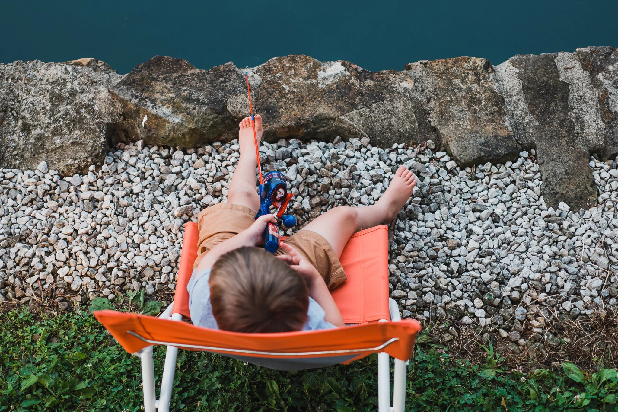 Child sitting on an orange lounge chair by the water, fishing with a rod, with rocks and green grass surrounding the area
