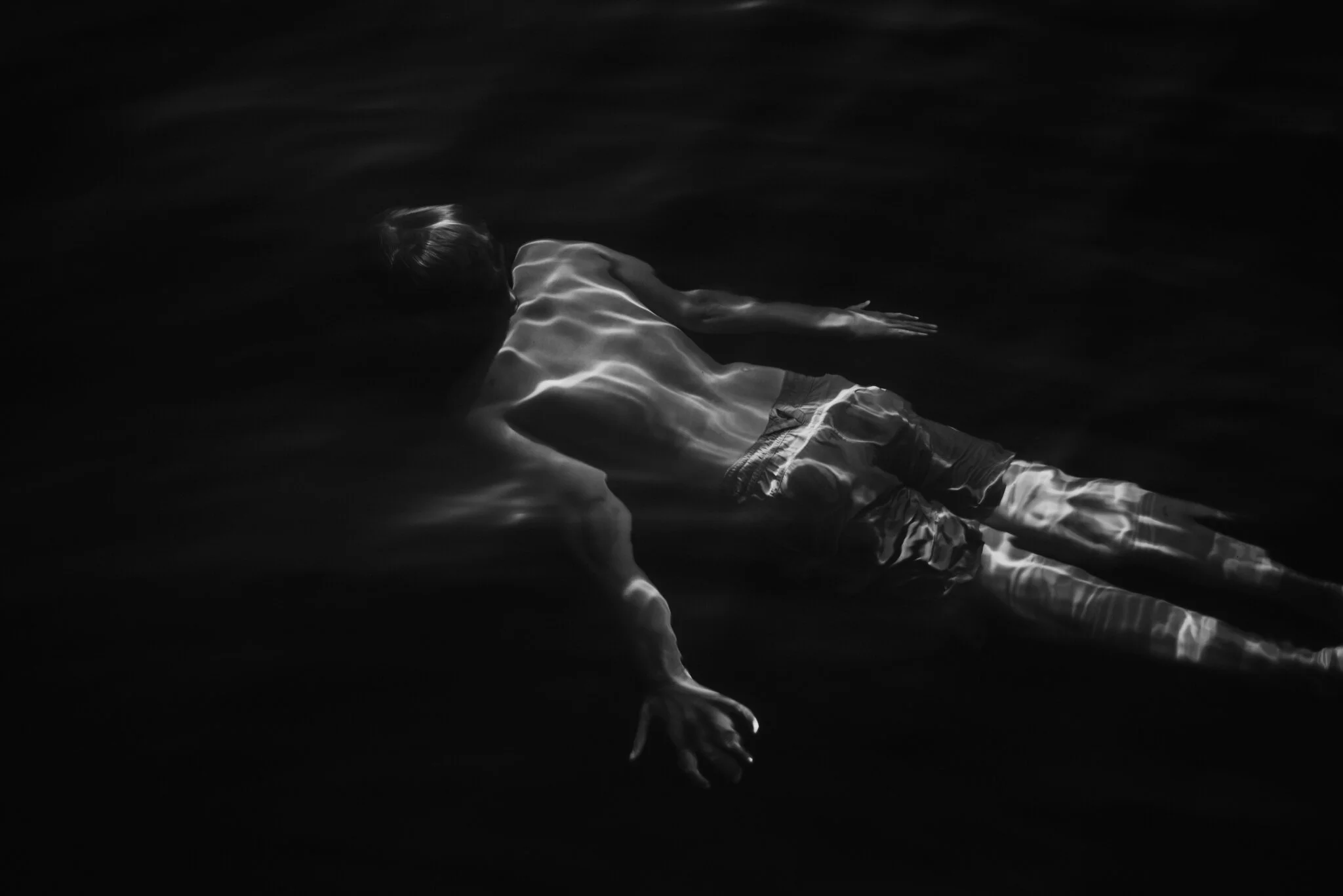 A person floating face down in dark water, with reflections creating light patterns on their body.