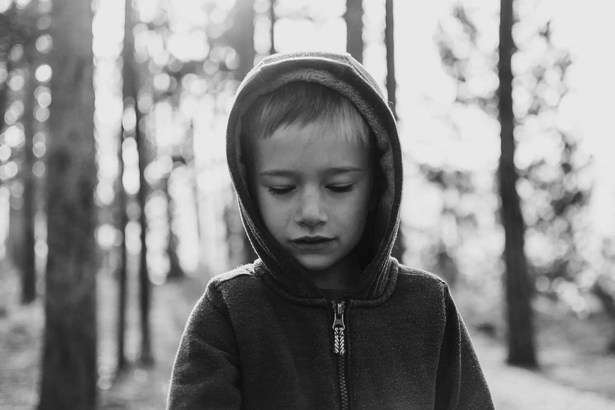 A young boy with closed eyes and a thoughtful expression, wearing a hooded jacket, standing in a forest with blurred trees in the background, black and white image.