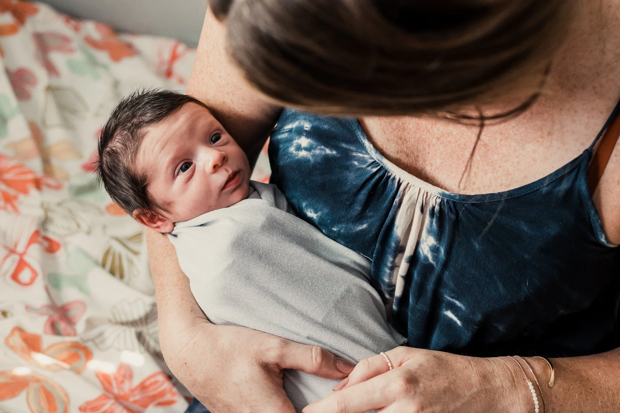 A woman holding a newborn baby close to her chest indoors, with a floral-patterned blanket in the background.