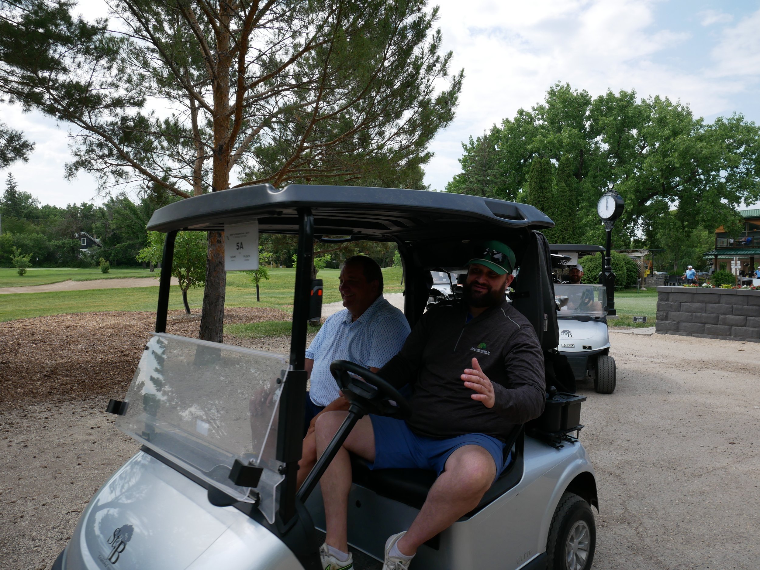 Two men sitting in a golf cart at a golf course, with trees, a clock, and a clubhouse in the background.