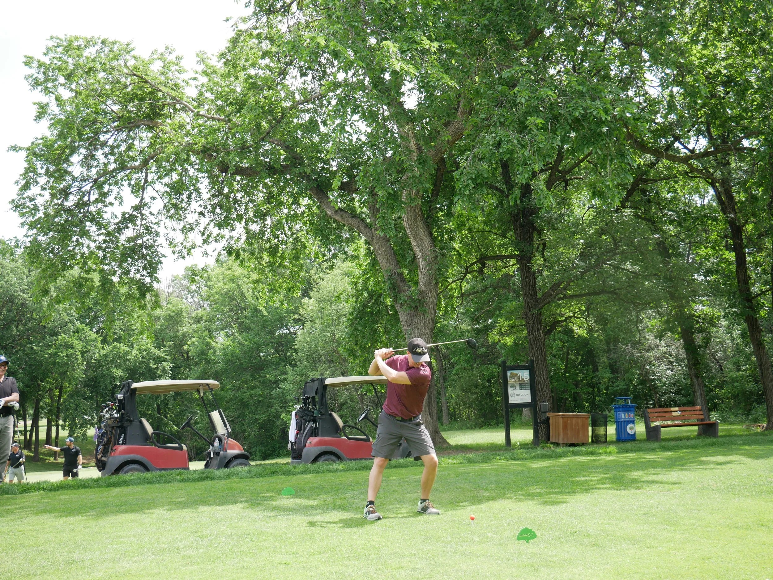 Person swinging a golf club on a golf course with golf carts and other people visible in the background.