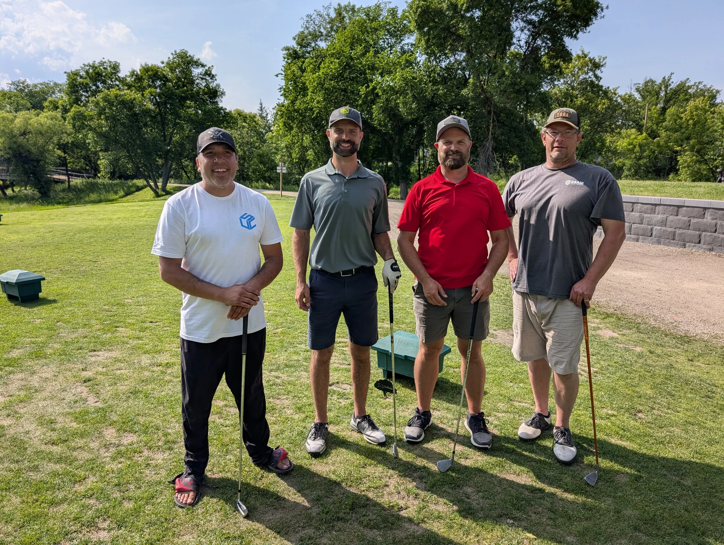 Four men standing on a golf course holding golf clubs, with trees and a clear blue sky in the background.