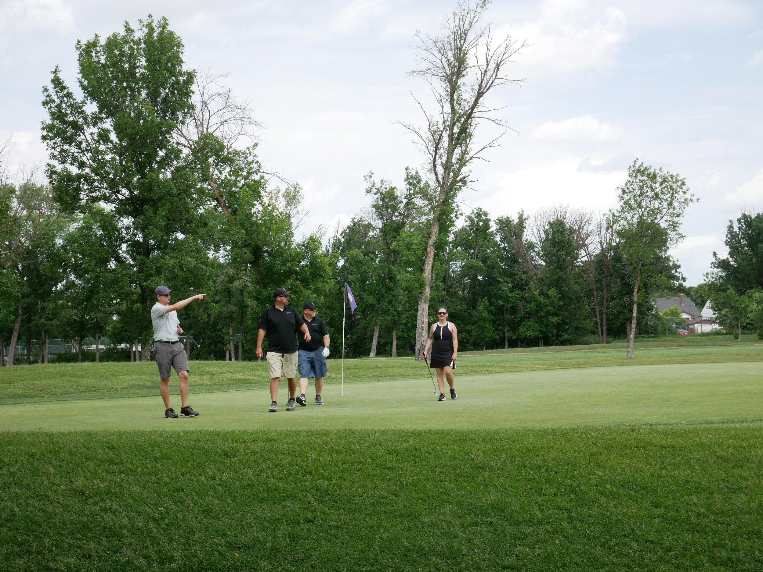 Four golfers on a golf course, with one person pointing, two people standing near the flag, and one woman walking towards the camera, surrounded by trees and houses in the background.