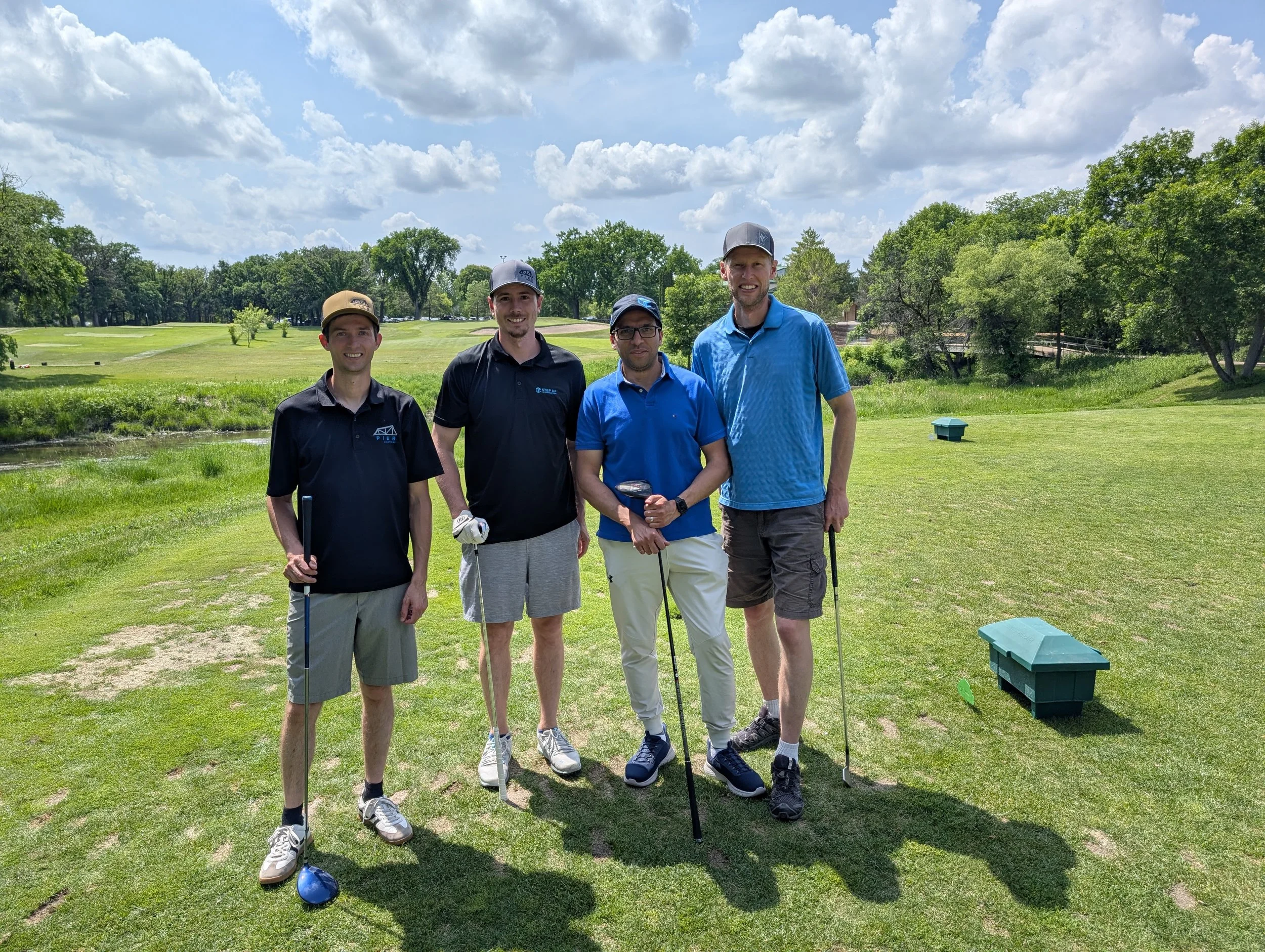 Four men standing on a golf course, smiling, with golf clubs, green grass, trees, blue sky with clouds in the background.