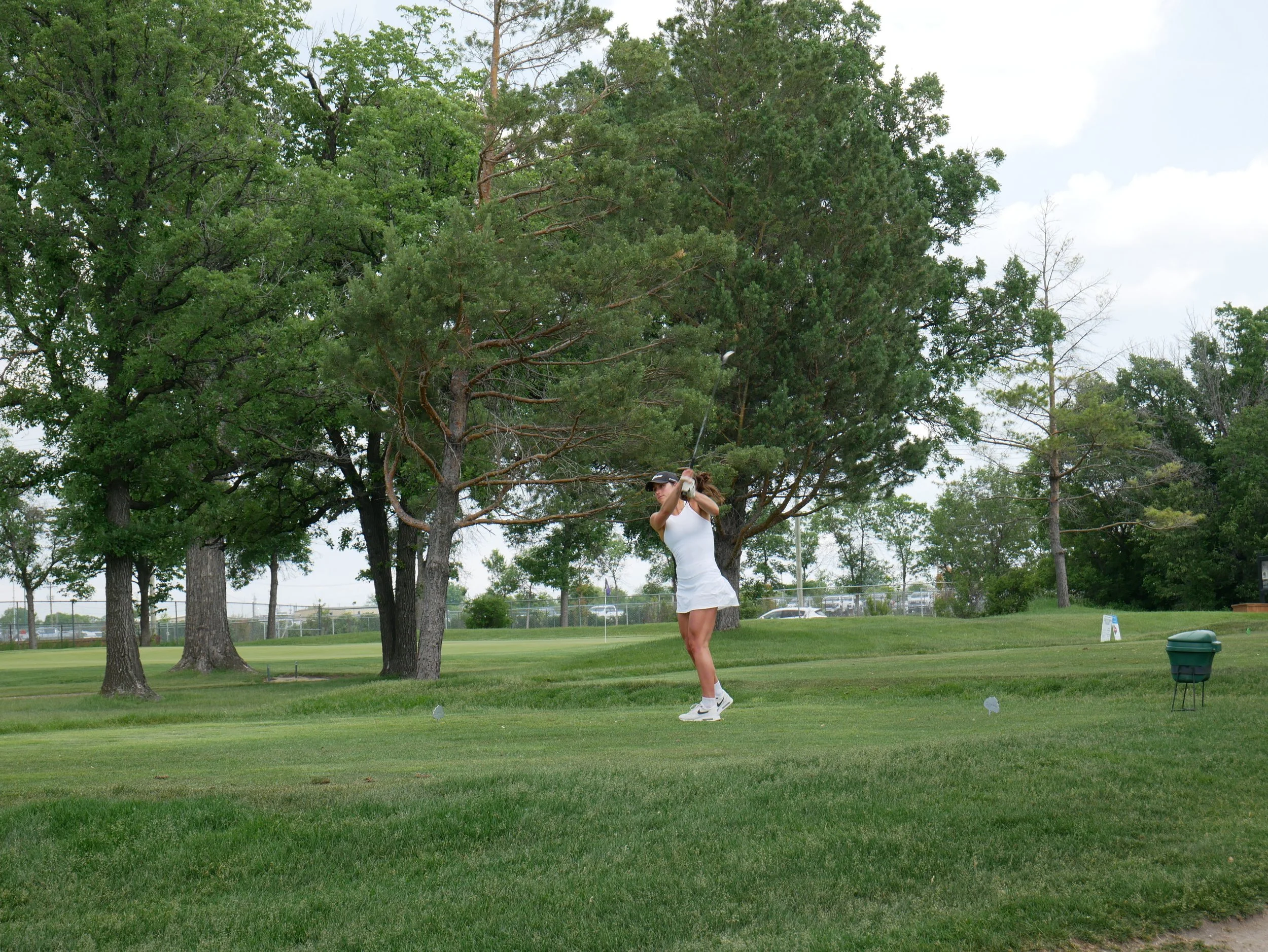 A woman in a white dress and cap playing golf on a course with green grass, trees, and a partly cloudy sky.