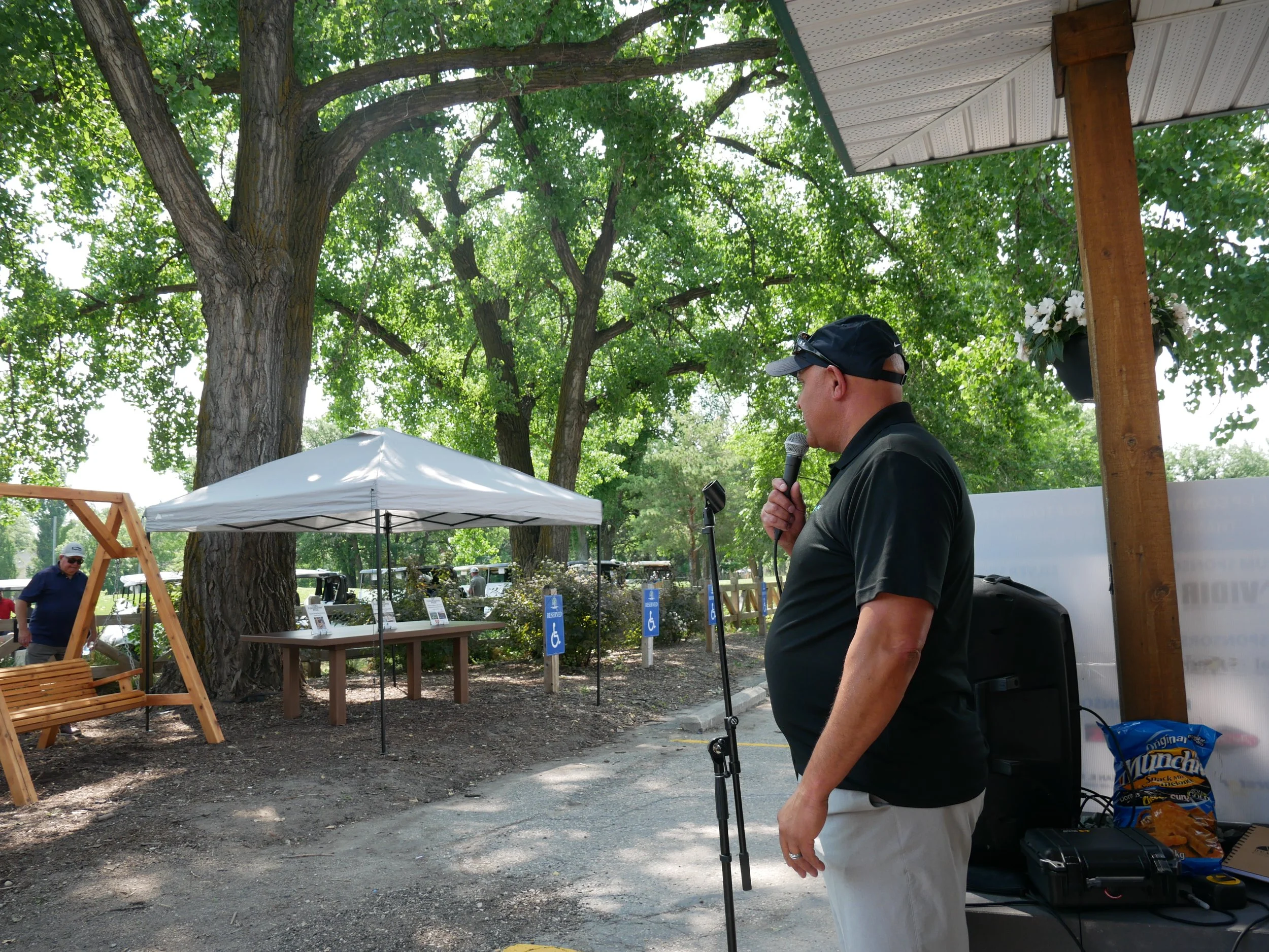A man in a black shirt and cap speaking into a microphone at an outdoor event shaded by large trees, with picnic tables, a white tent, and people in the background.
