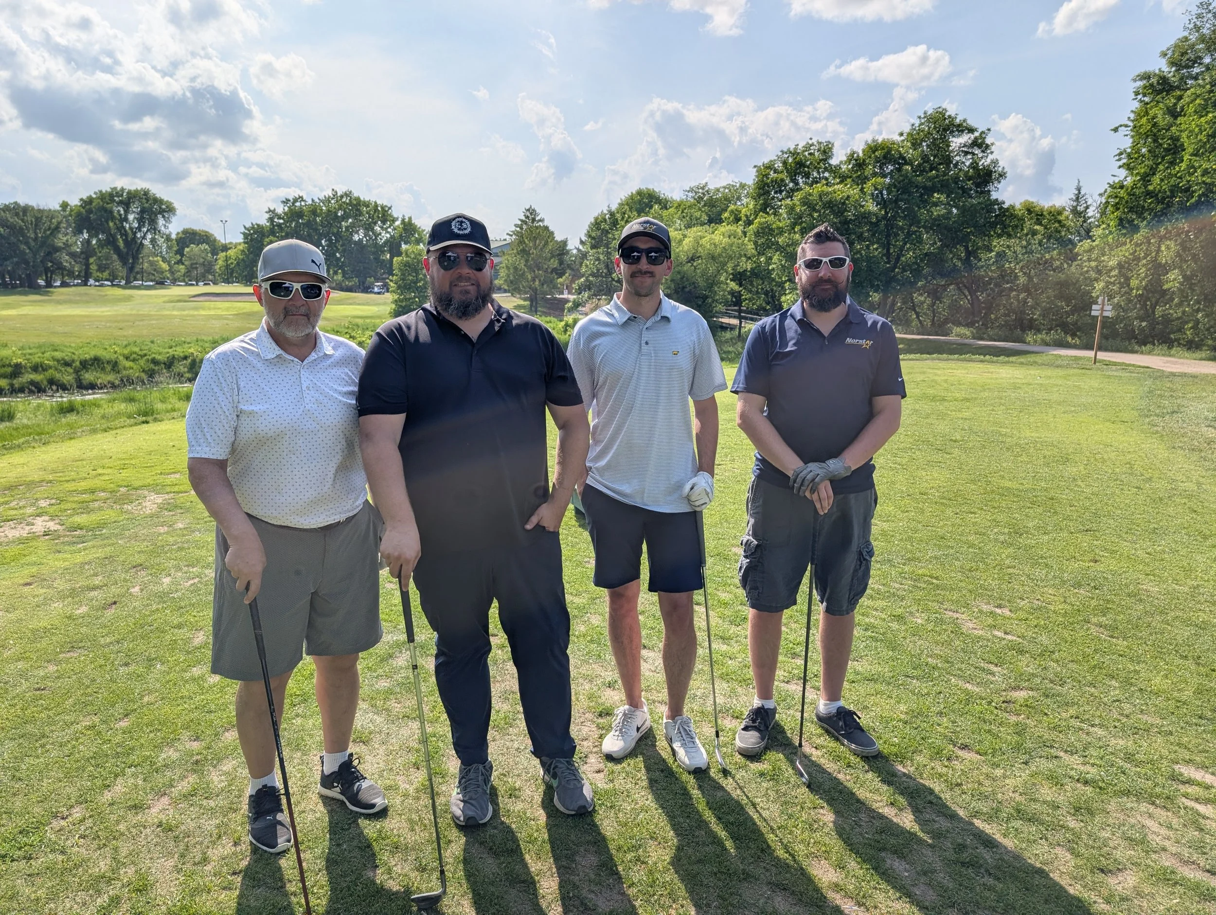 Four men standing on a golf course holding golf clubs on a sunny day with trees and blue sky in the background.