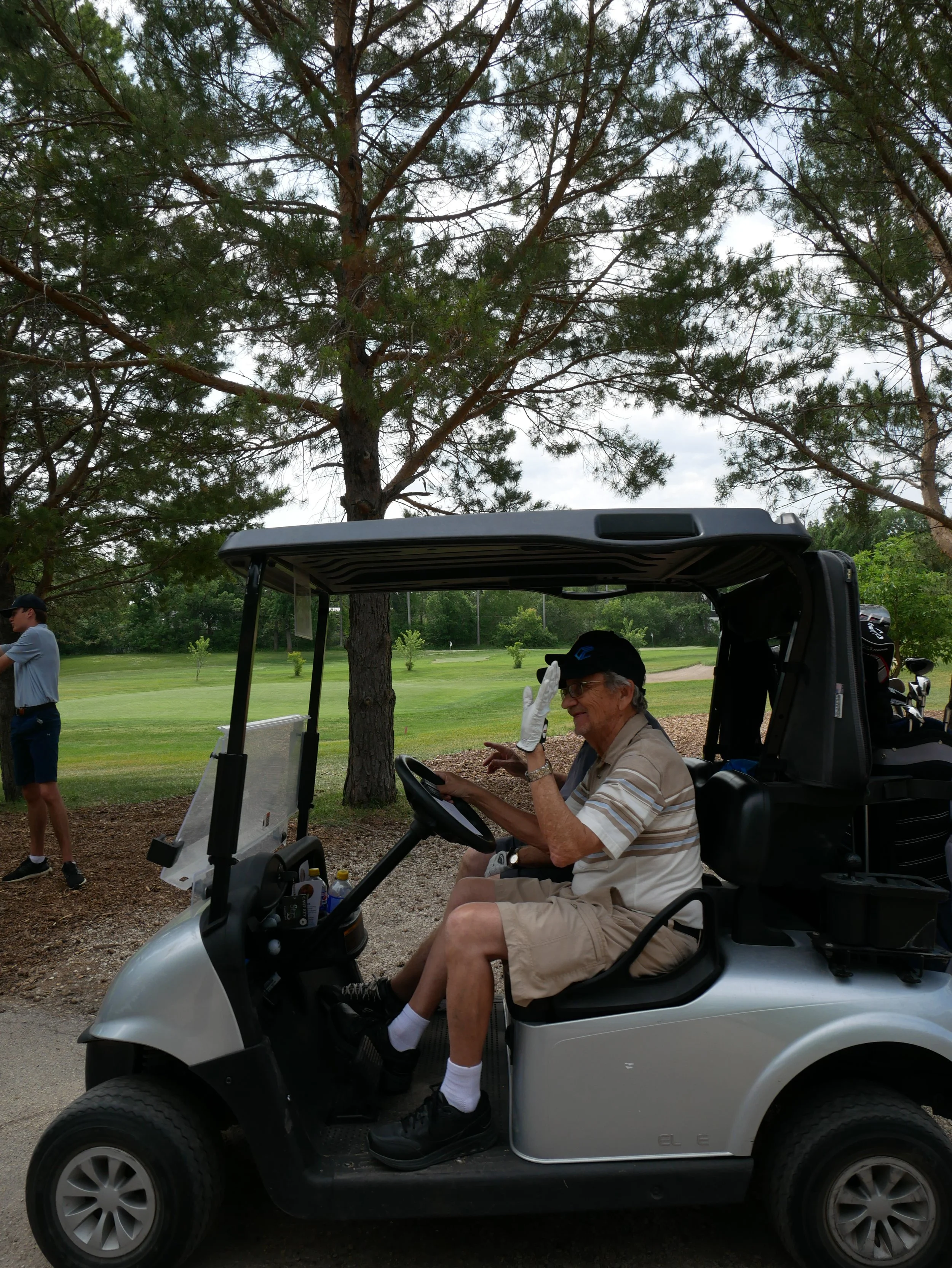 An elderly man in a golf cart on a golf course, waving and smiling.