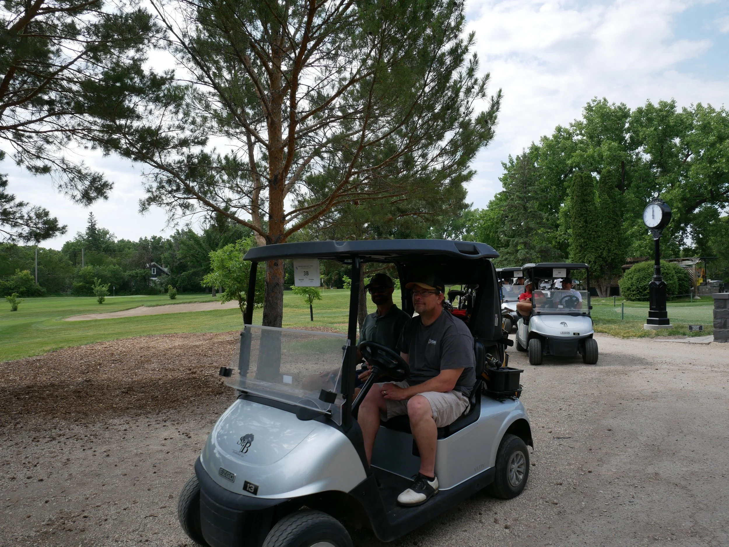 Two men sitting in a golf cart on a golf course, with trees, a clock, and another golf cart in the background.