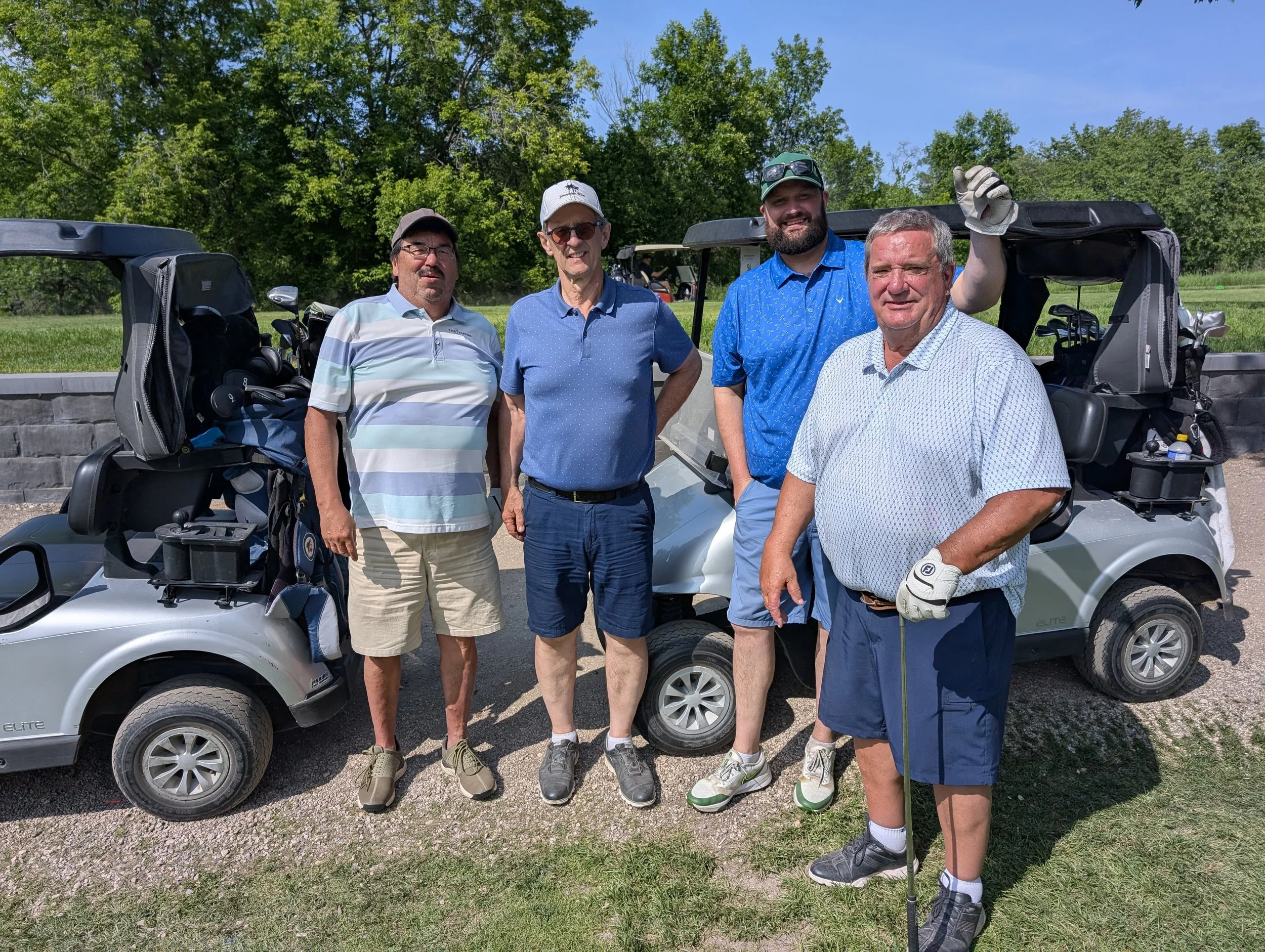 Five men standing on a golf course beside two golf carts with golf bags, smiling at the camera on a sunny day.