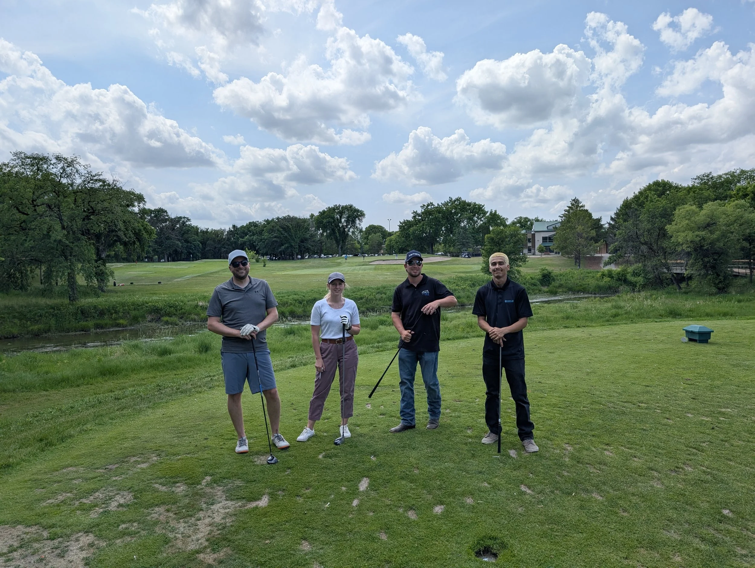 Four people standing on a golf course holding golf clubs, smiling, with trees and a partly cloudy sky in the background.