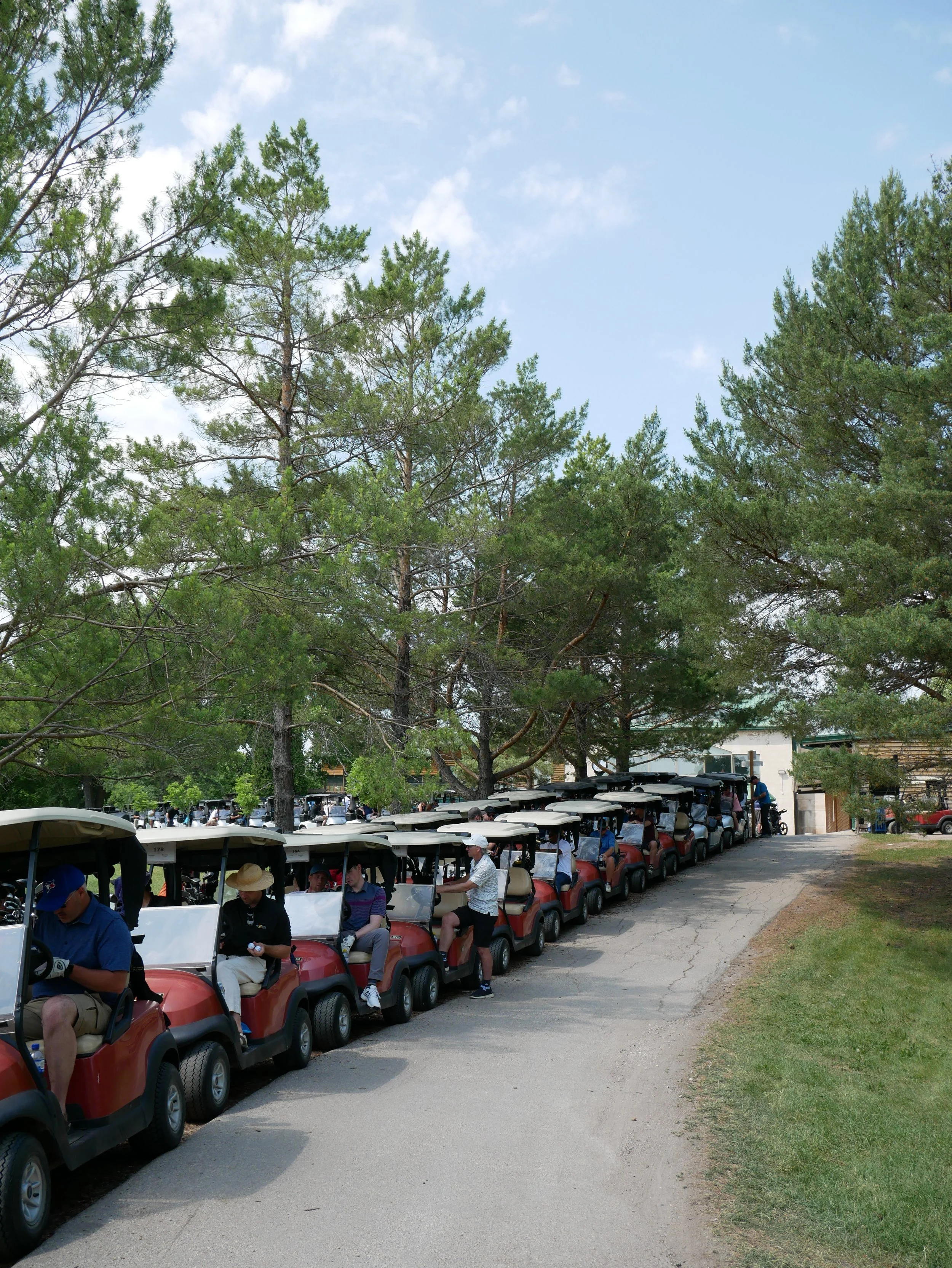 Line of parked golf carts under trees on a golf course with people sitting and preparing to play.