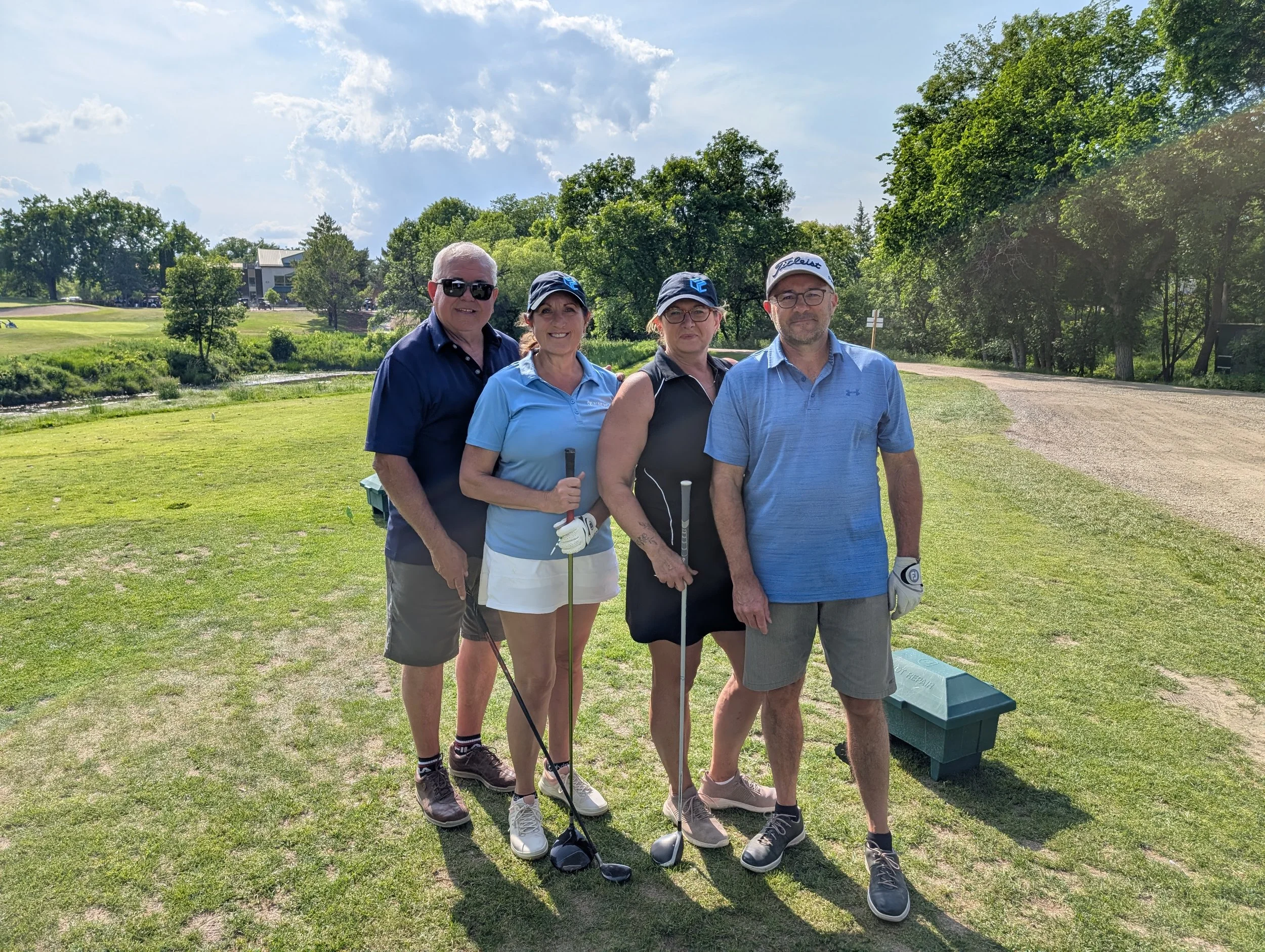 Four people standing on a golf course, posing for a photo. They are dressed in golf attire, holding golf clubs, with a sunny sky and trees in the background.