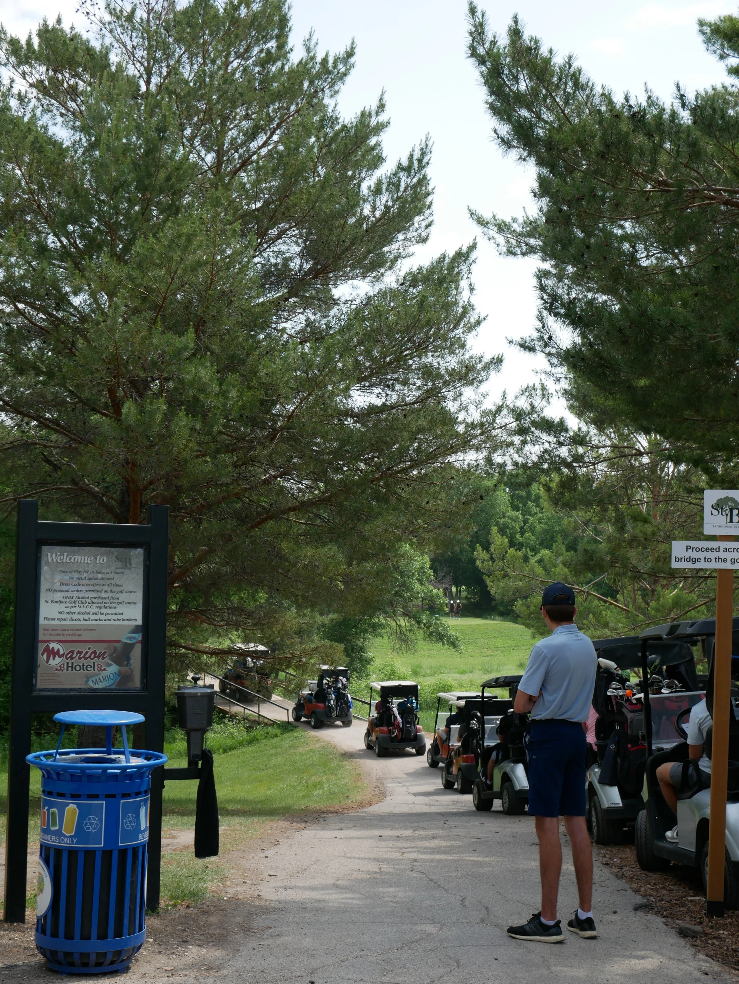 A man standing near golf carts on a golf course path, surrounded by trees with a page of rules and instructions posted nearby.