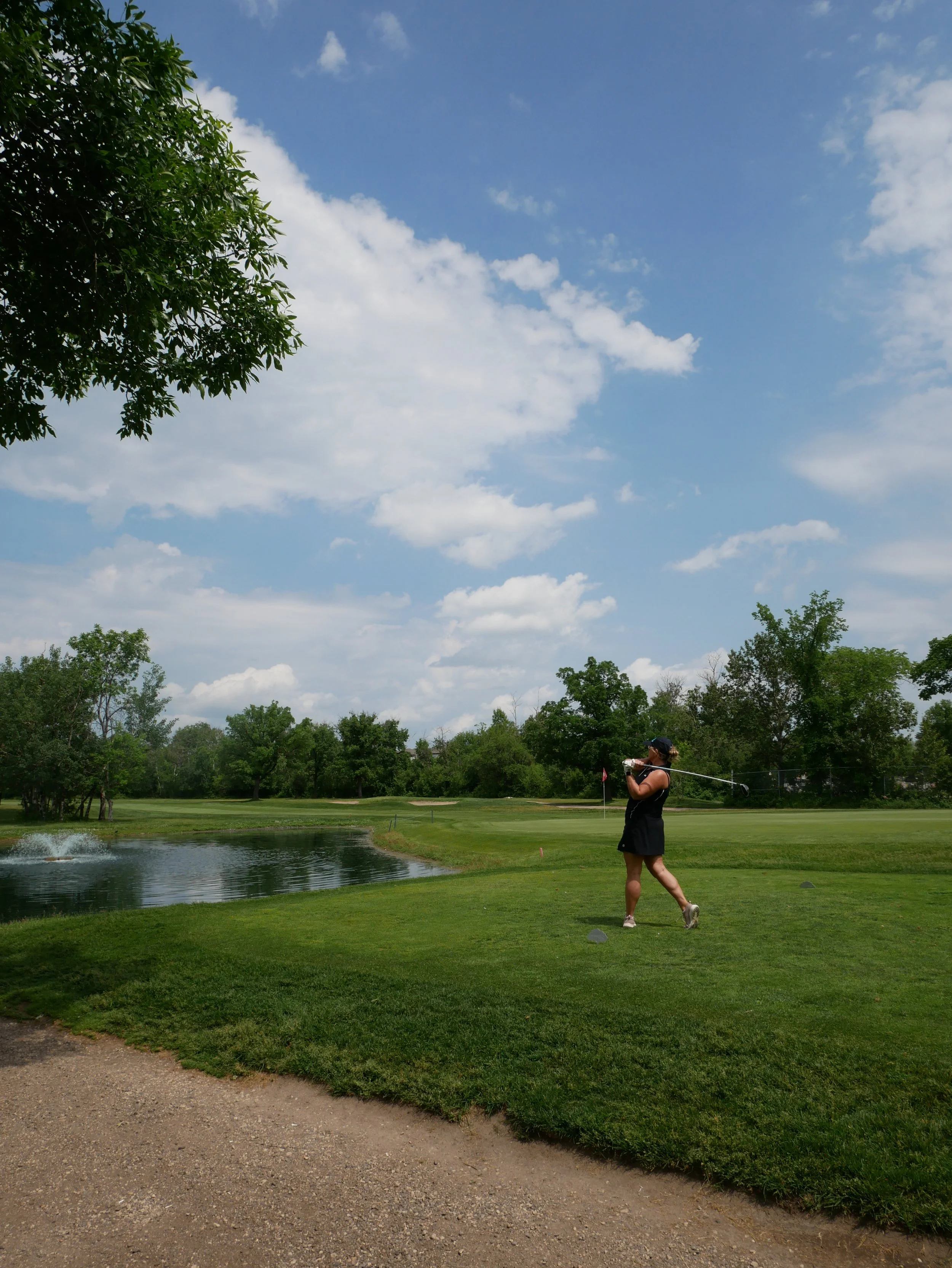A woman in a black dress and cap swinging a golf club on a golf course near a water hazard with trees and blue sky with clouds in the background.