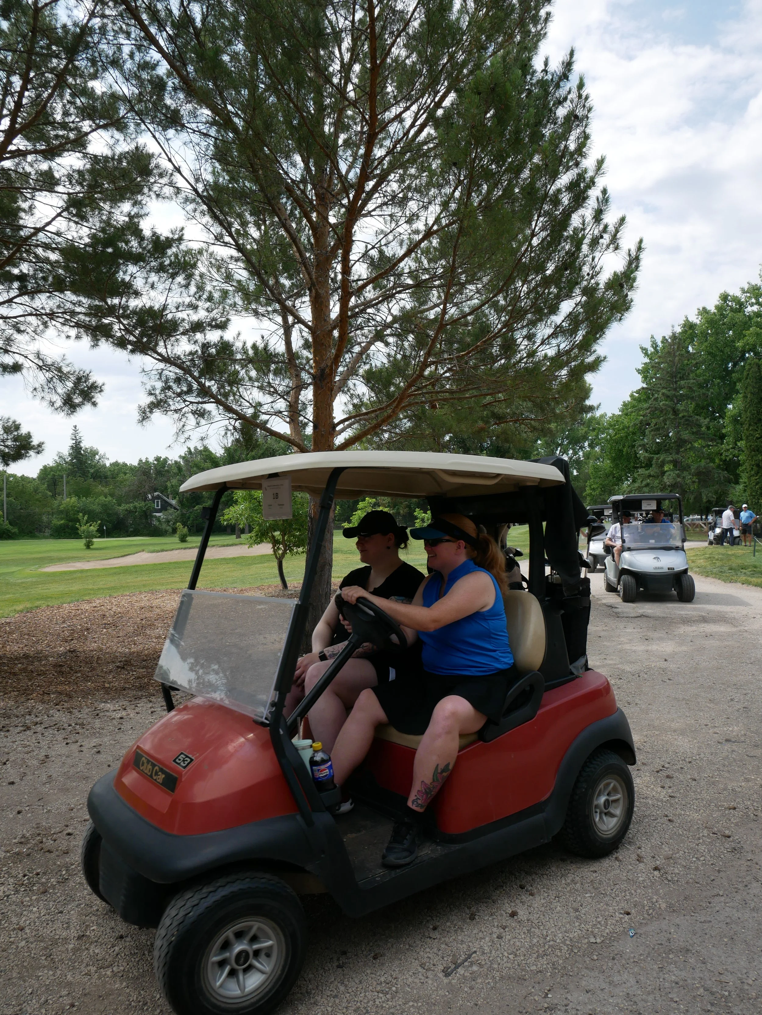 Two women sitting in a red golf cart on a golf course, with trees and a person driving another golf cart in the background.