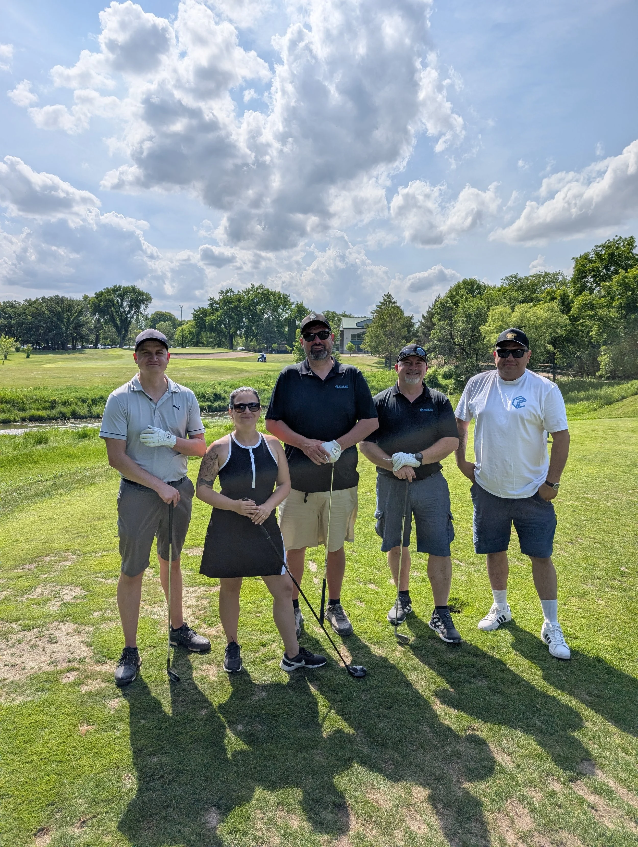 Five people standing on a golf course holding golf clubs, posing for a photo under partly cloudy sky.