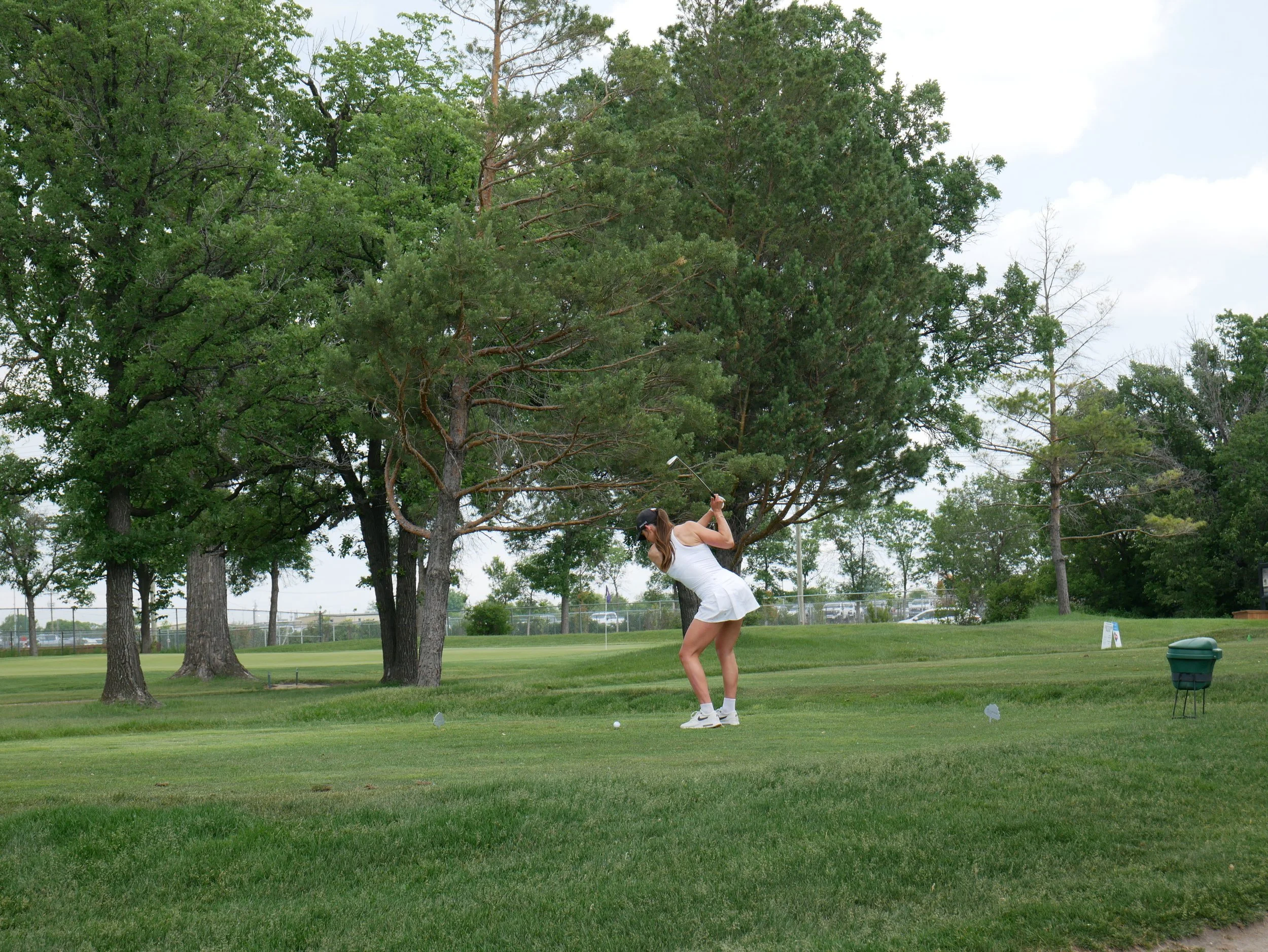 A woman playing golf, swinging a club on a golf course with green grass, trees, and cloudy sky.