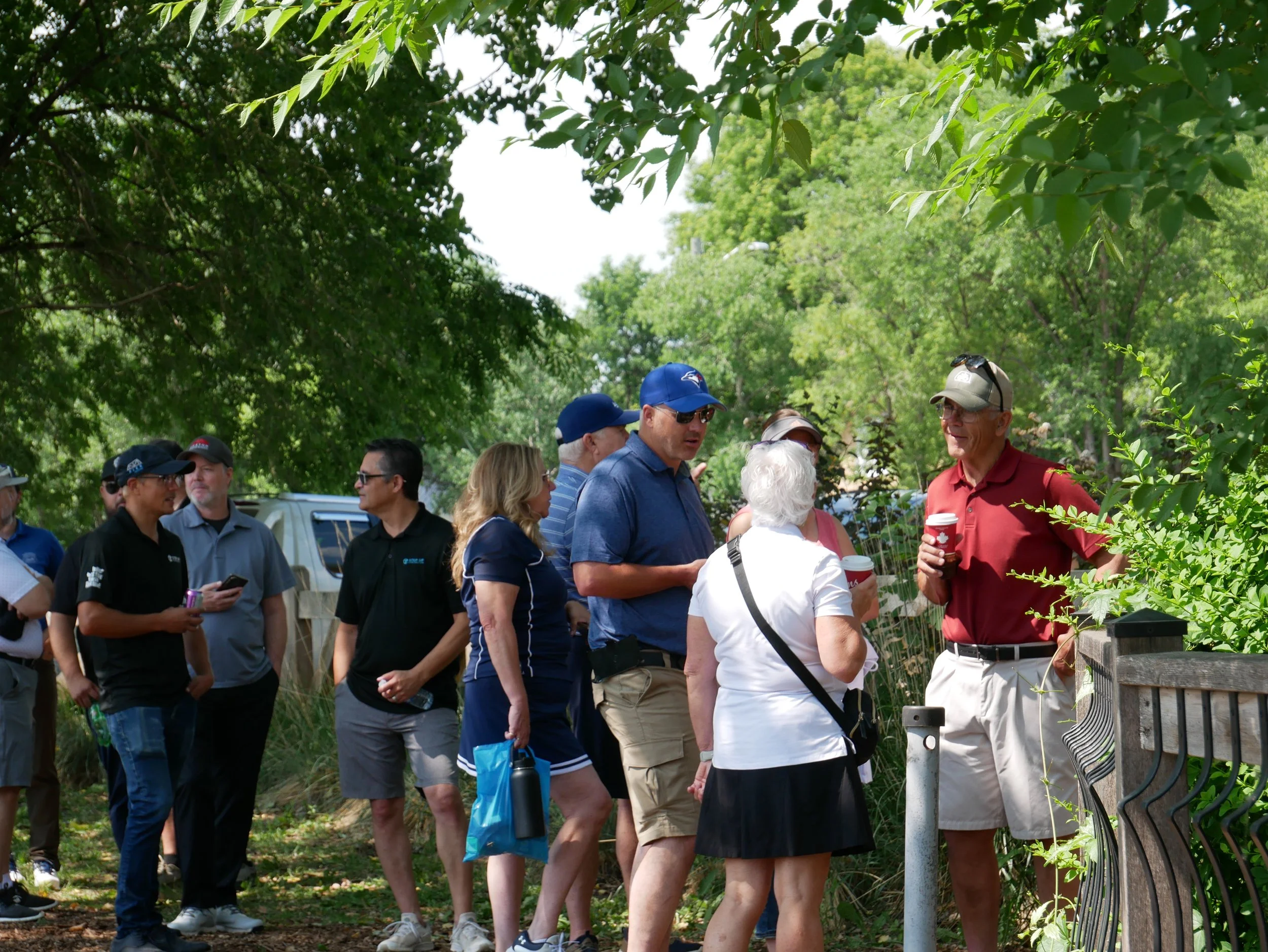 A group of people standing outdoors near a fence, engaging in conversation on a sunny day surrounded by green trees.
