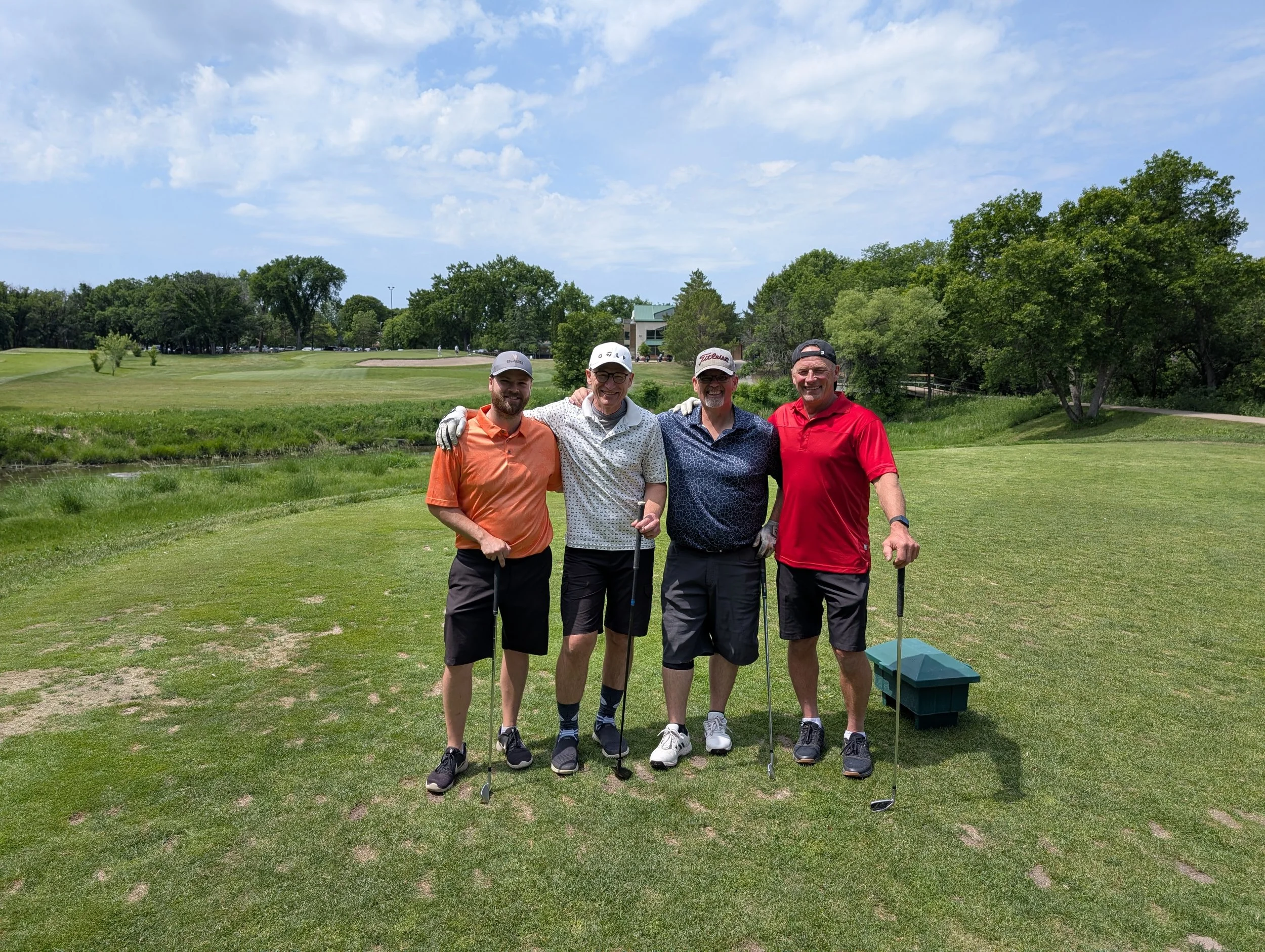 Four men standing on a golf course with golf clubs, smiling for a photo. They are dressed in golf attire and are standing on the green near a sand trap, with trees, a pond, and a building in the background. The sky is partly cloudy.
