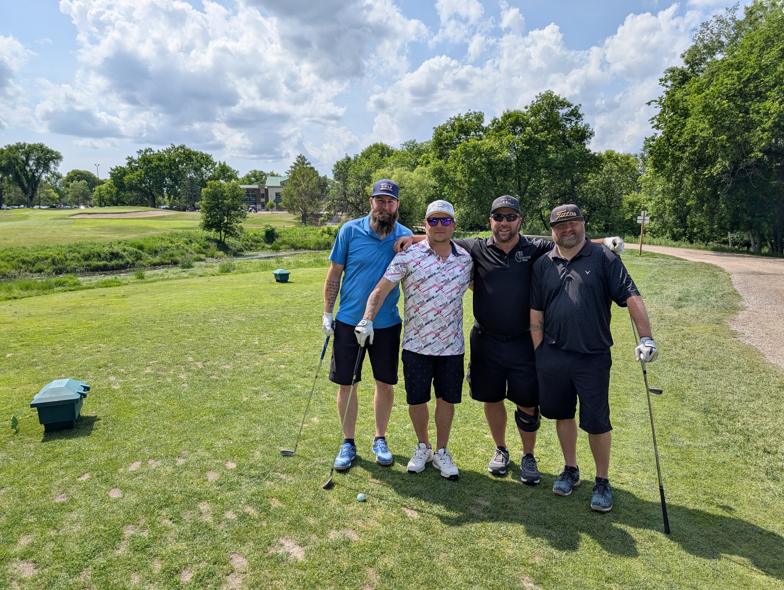 Four men standing together on a golf course, smiling, with golf clubs in hand. They are dressed in golf attire, and the background shows trees, a building, and a cloudy sky.