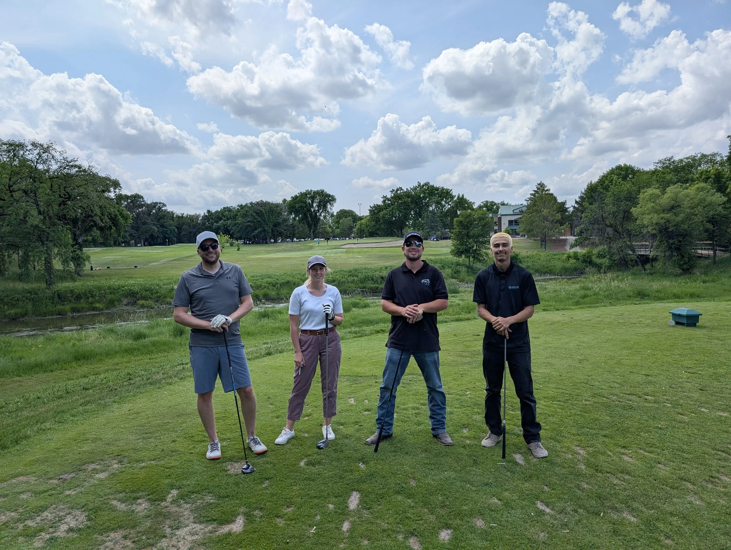 Four people standing on a golf course holding golf clubs, with trees, green grass, and a partly cloudy sky in the background.