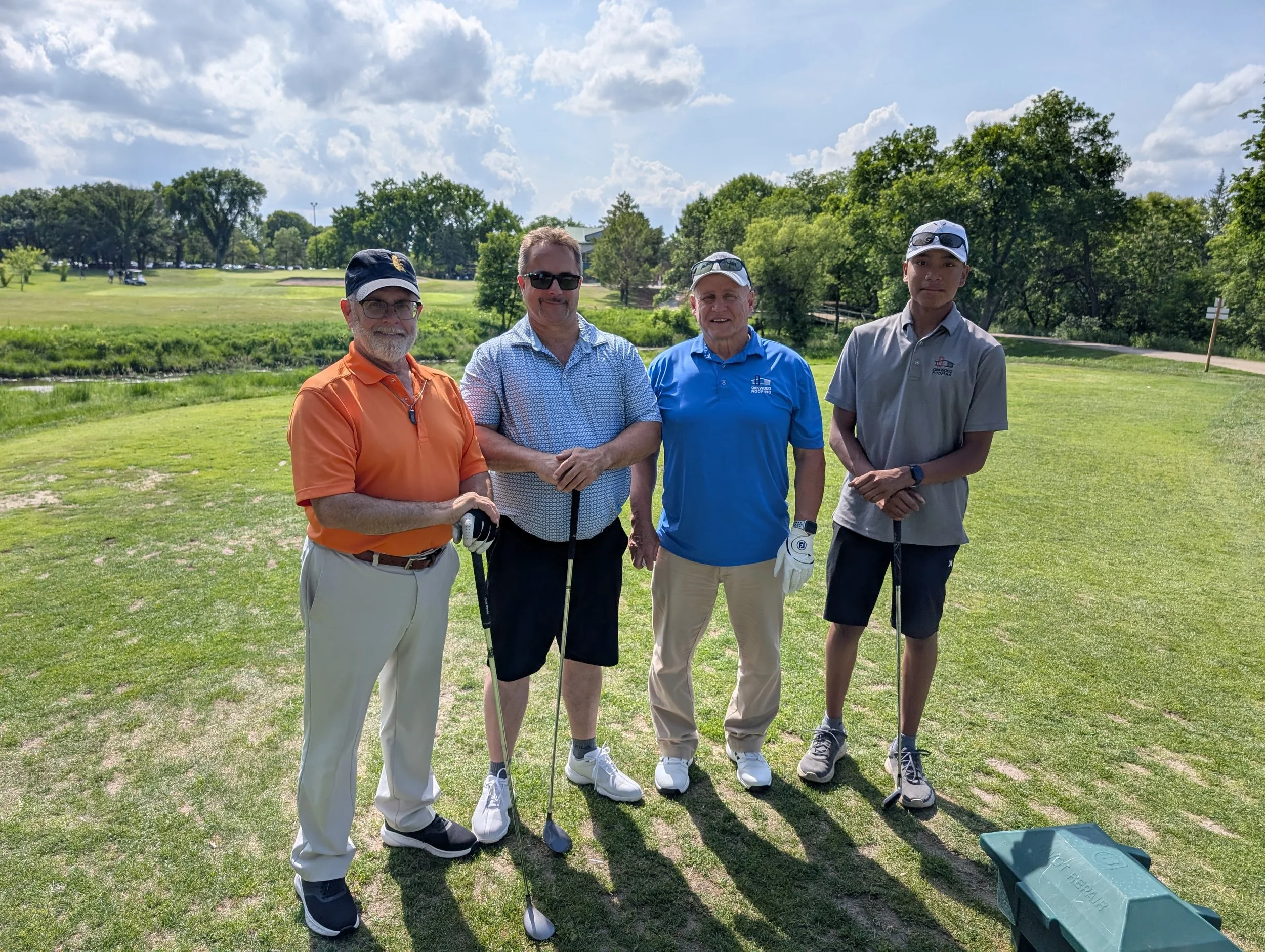 Four men standing on a golf course holding golf clubs, with trees and a partly cloudy sky in the background.