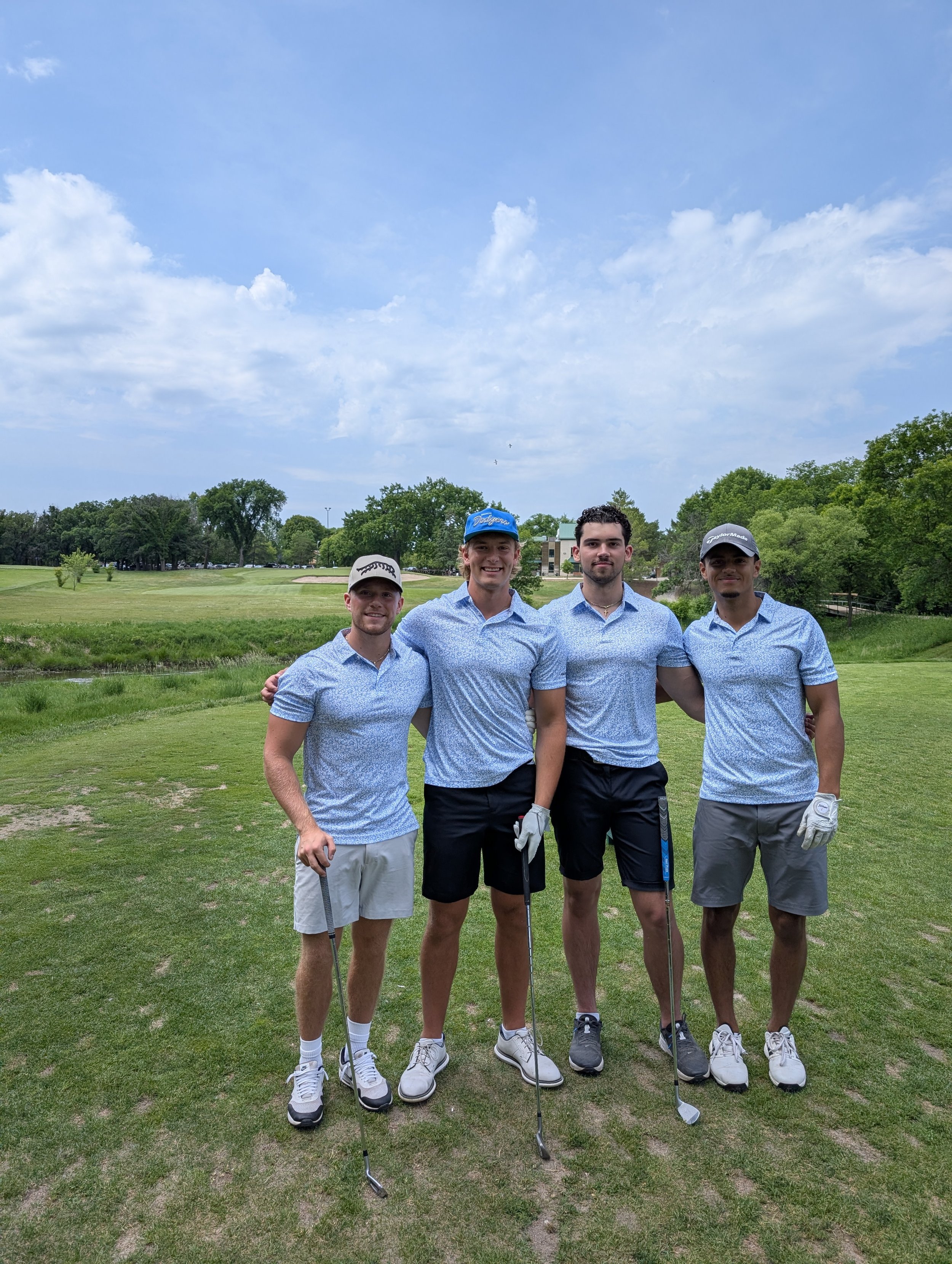 Four young men in golf attire standing on a golf course with trees and a blue sky in the background.