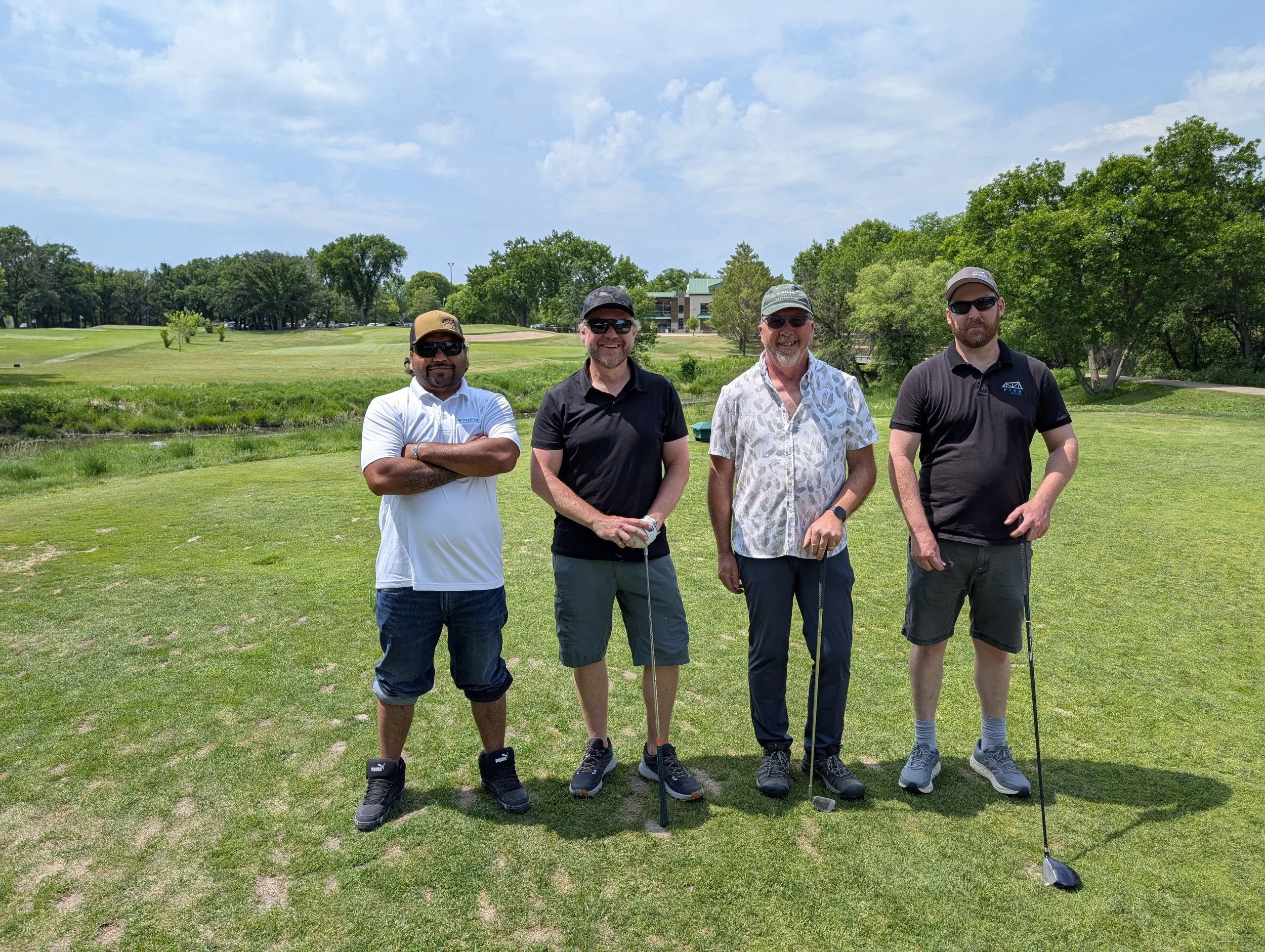 Four men standing on a golf course holding golf clubs, smiling, under a partly cloudy sky with trees and a building in the background.