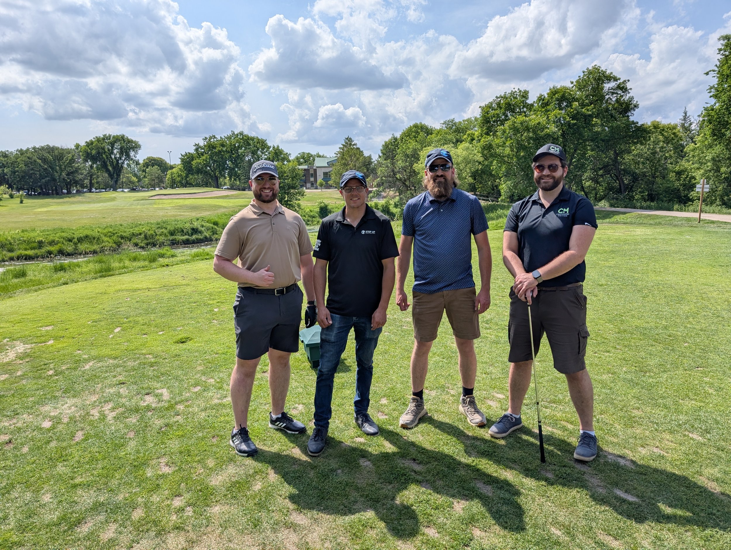 Four men standing on a golf course, dressed in golf attire, with trees and a cloudy sky in the background.