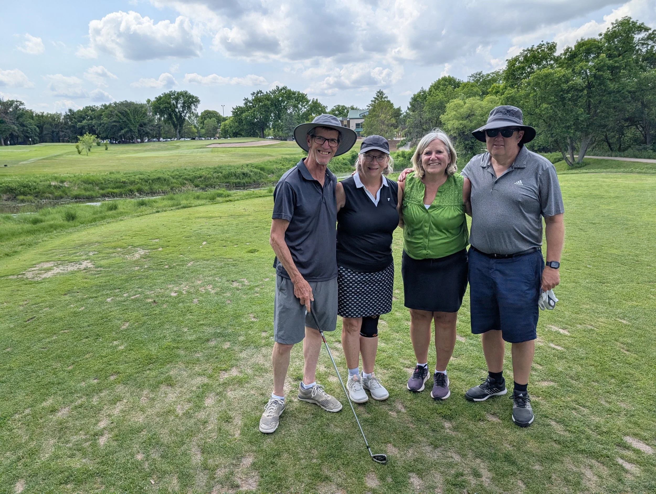 Four people standing on a golf course, smiling and posing for a photo. Two men and two women, dressed in golf attire, with trees and a putting green in the background under a partly cloudy sky.