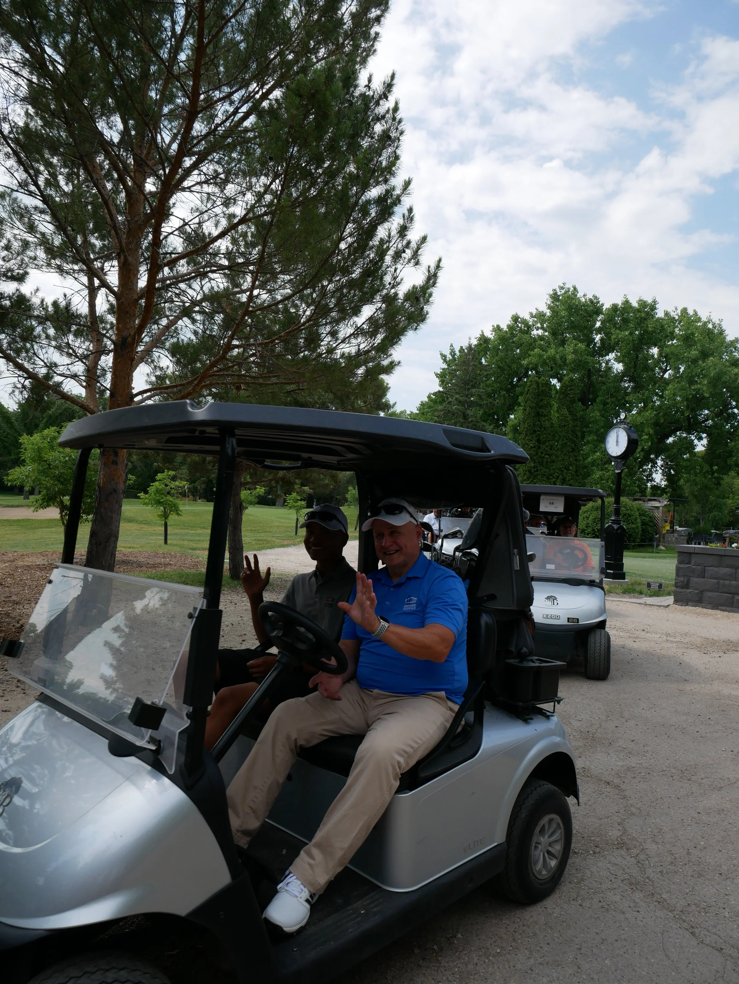 Two men riding in a golf cart, waving and smiling, on a golf course with trees in the background.