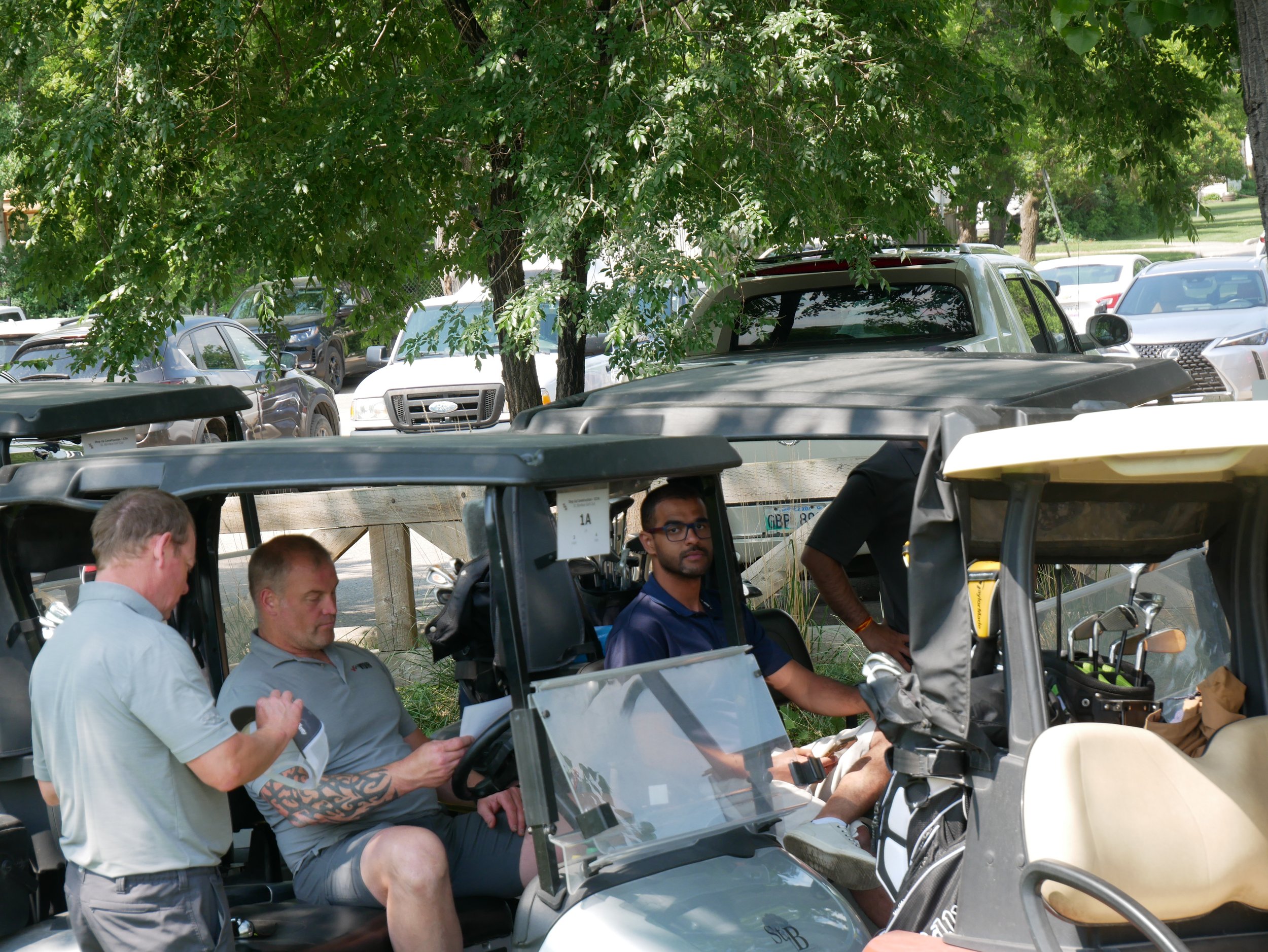 Three men sitting and standing around a golf cart on a sunny day, with parked cars and trees in the background.