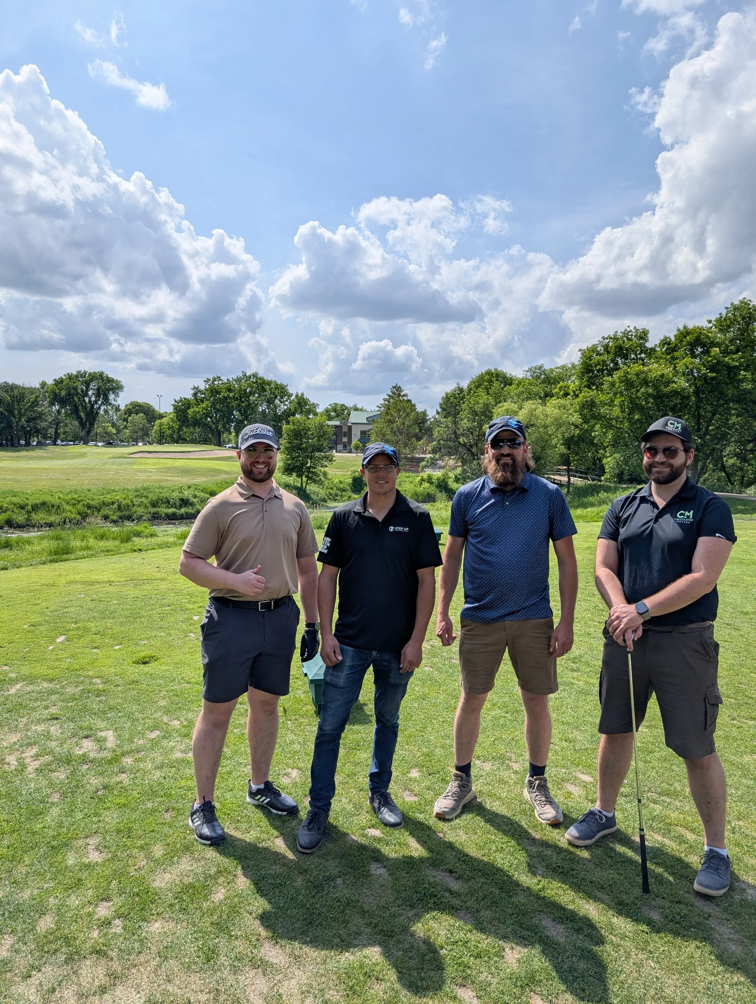 Four men standing on a golf course under a partly cloudy sky, dressed in golfing attire, with a golf club in hand of one of the men.