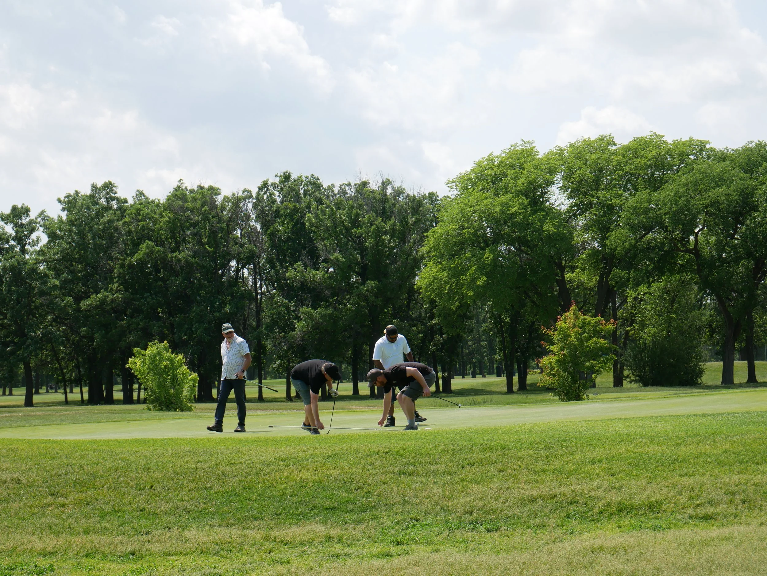 Four men playing golf on a sunny day, preparing to putt on the green with trees and blue sky in the background.