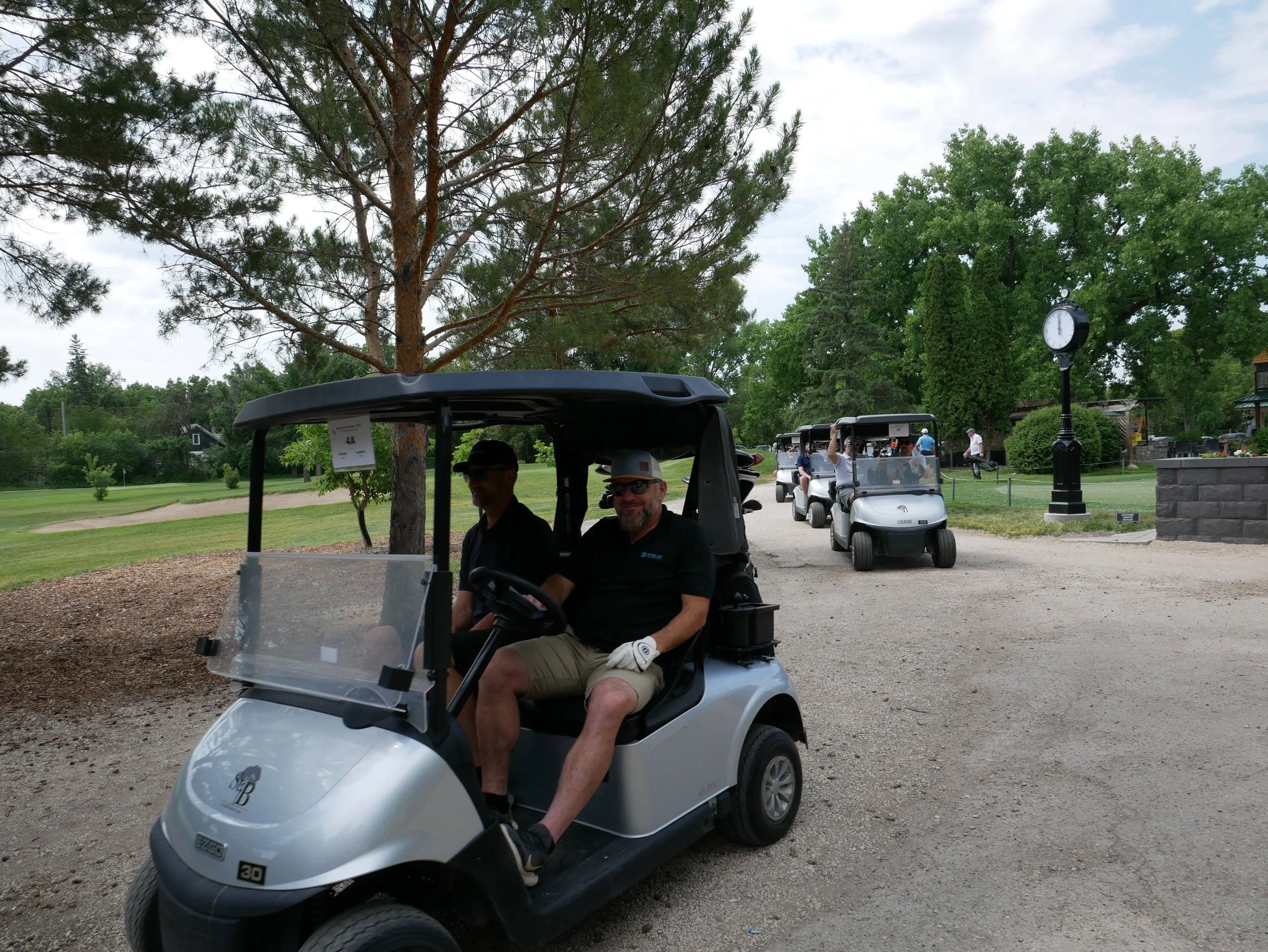Two men sitting in a golf cart on a golf course with multiple golf carts behind them, a large clock on a post, and surrounded by green trees and a cloudy sky.