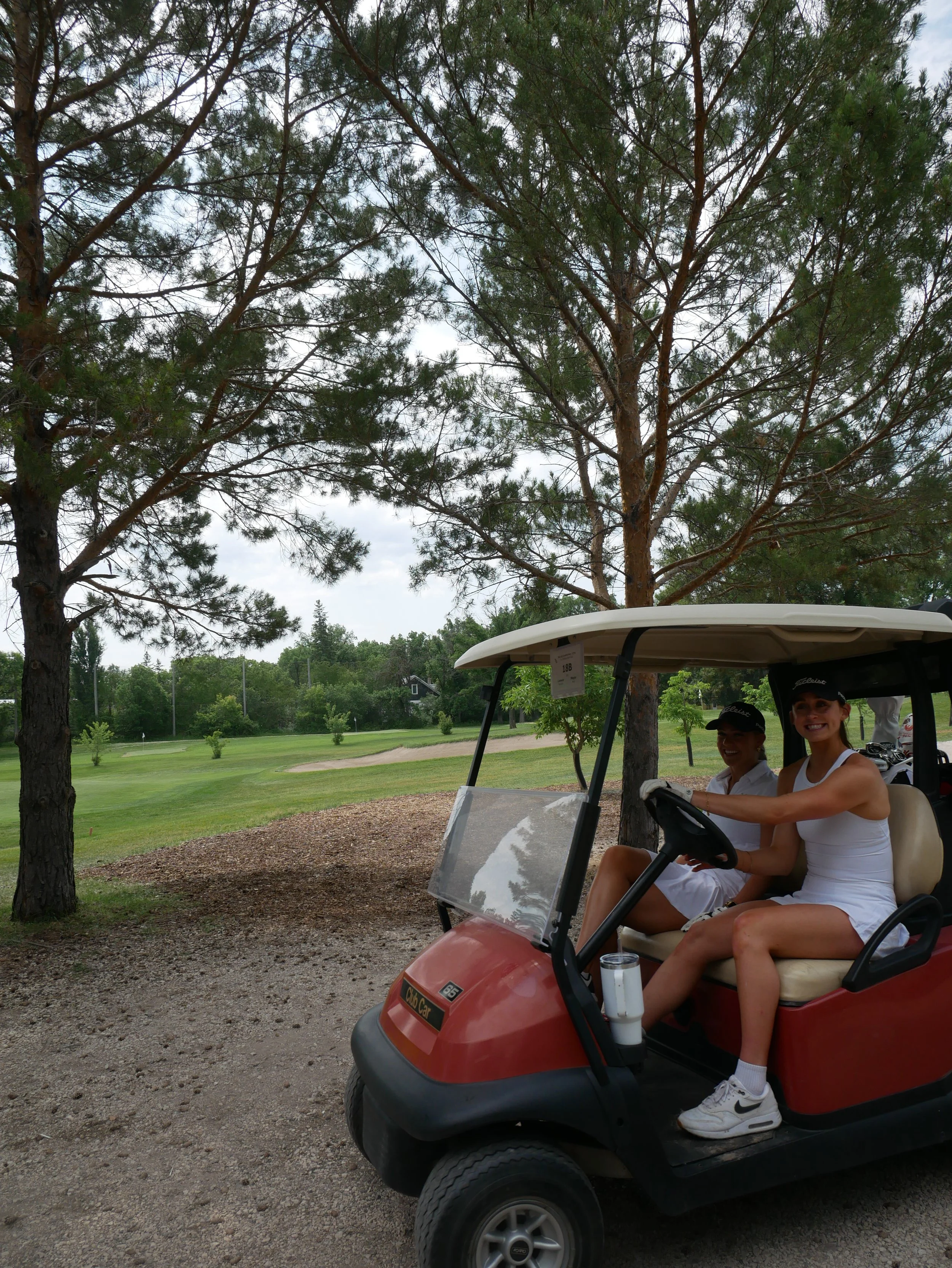 Two women in white golf outfits sitting in a red golf cart on a golf course with trees and green grass in the background.