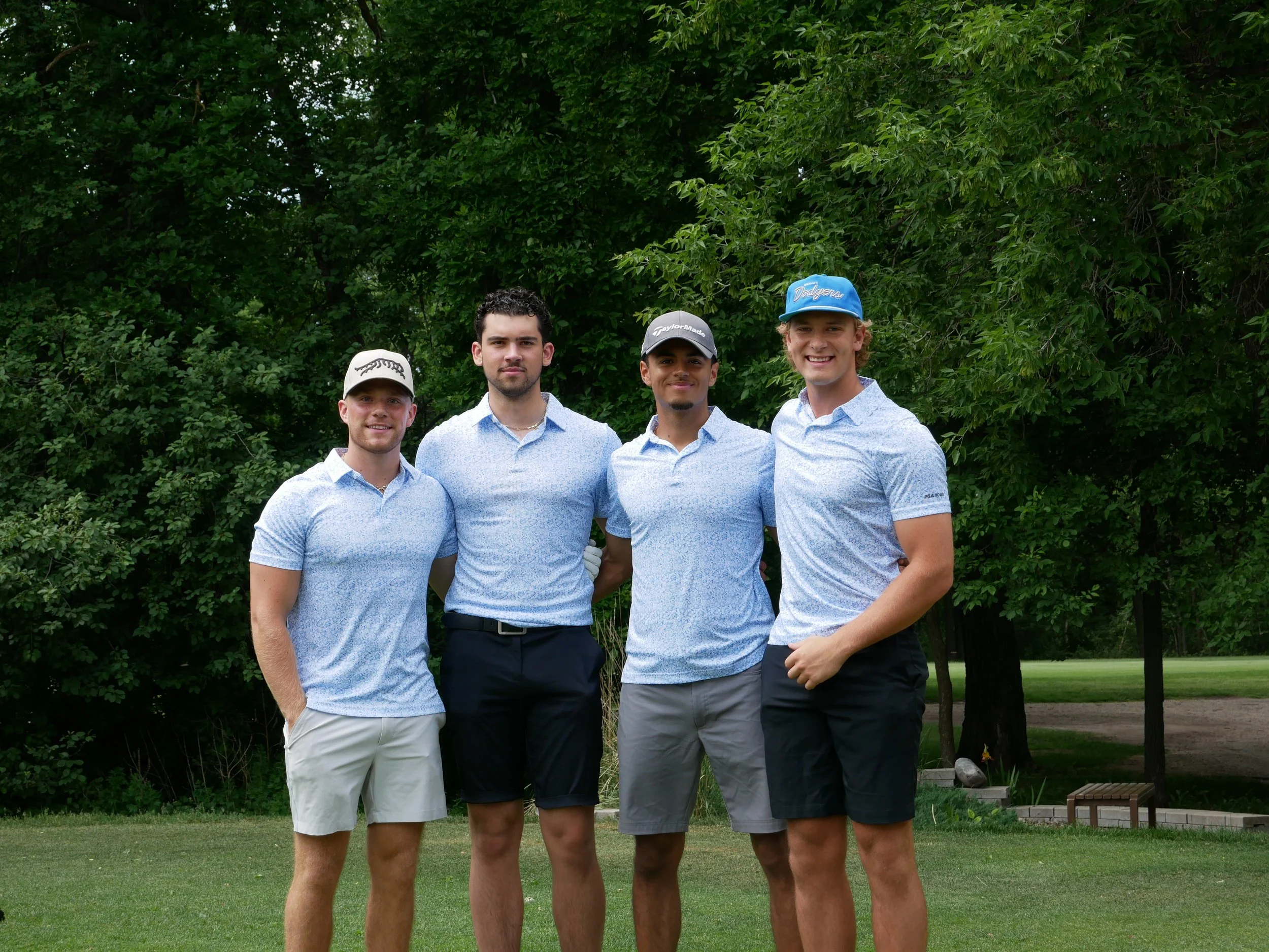 Four young men standing on a golf course, dressed in golf attire, with trees in the background.