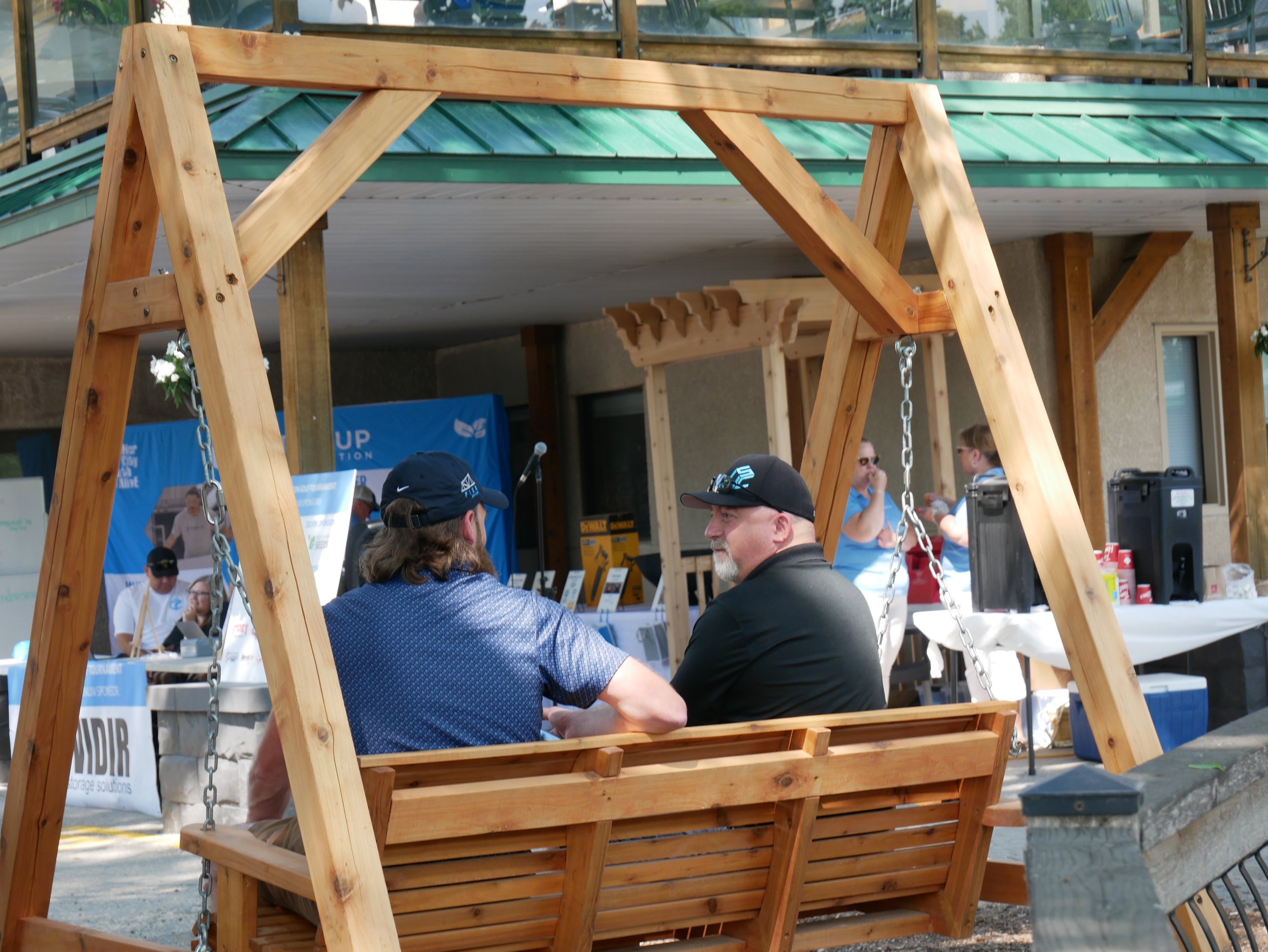 Two men sitting on a wooden swing bench, engaging in conversation, at an outdoor event with booths and banners in the background.