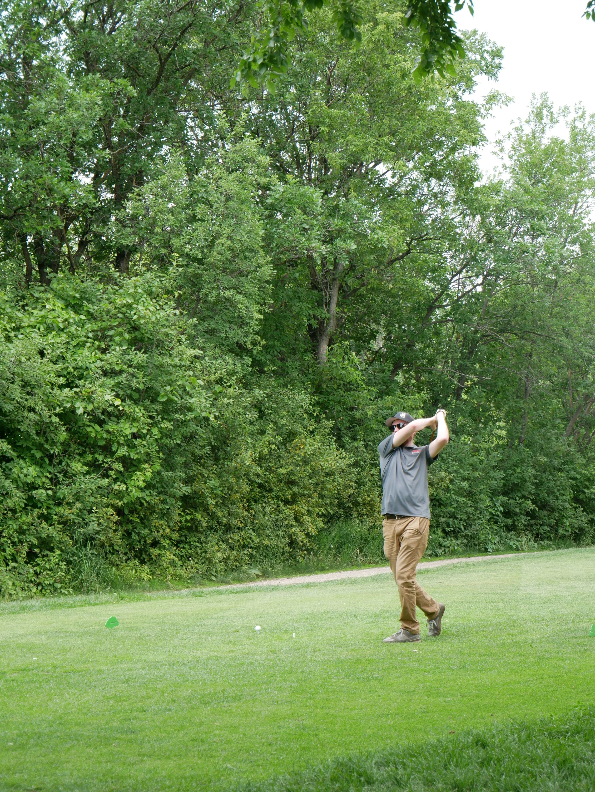 A man playing golf on a grassy course with trees in the background, swinging a golf club.