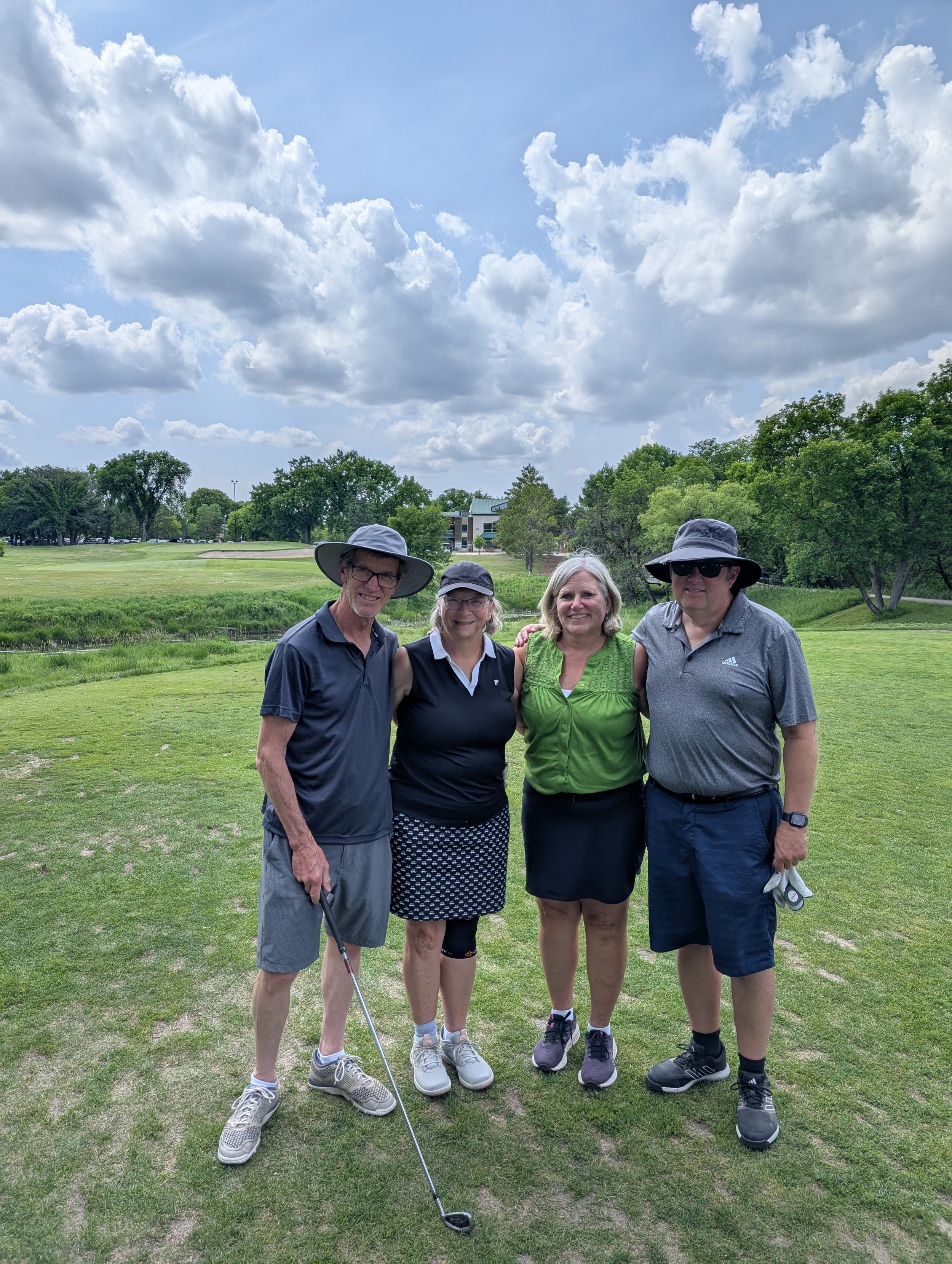 A group of four people standing on a golf course, smiling, with trees and a partly cloudy sky in the background.